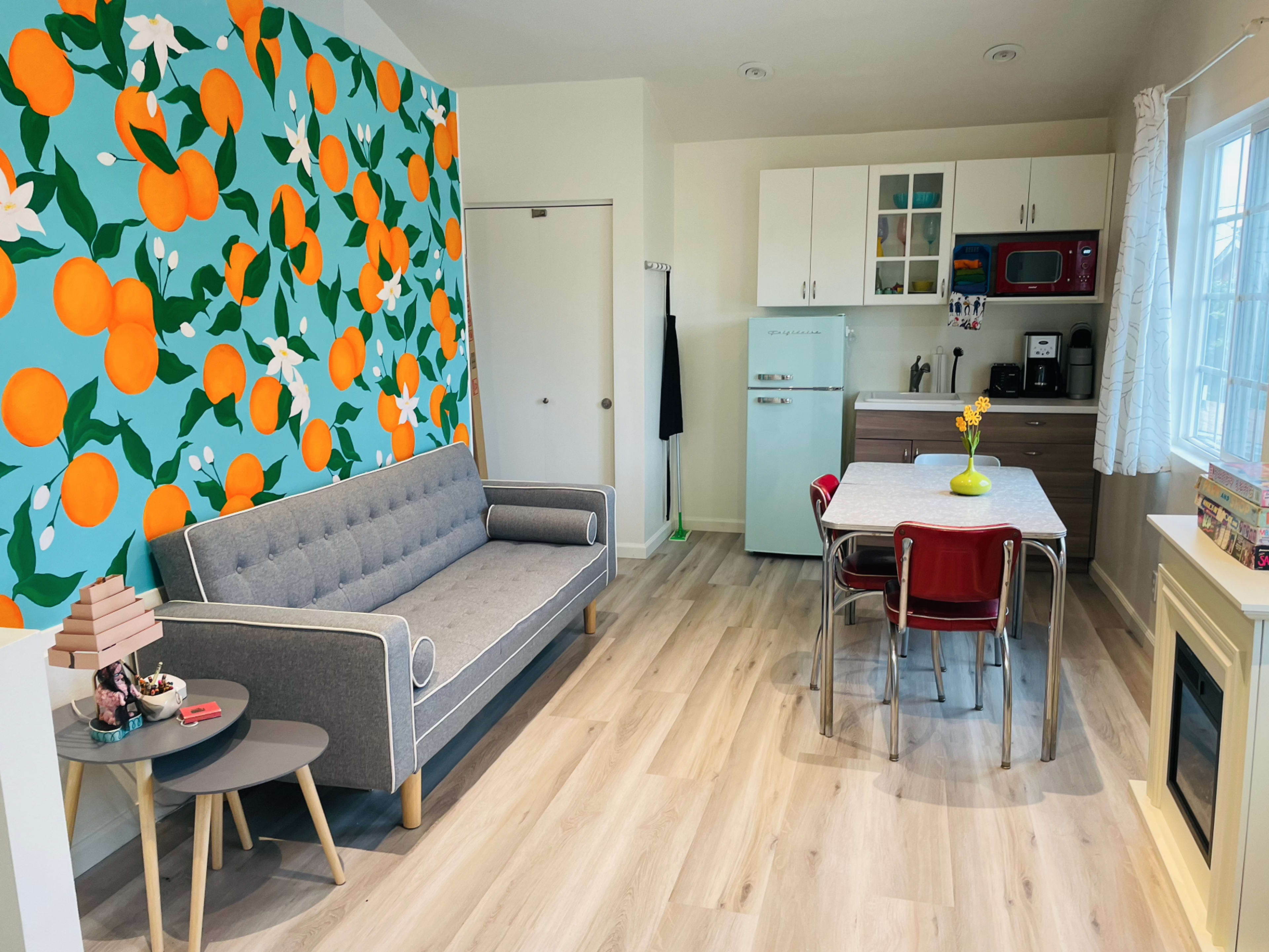 A small living space featuring a gray sofa, a patterned orange wall, a kitchenette with modern appliances, a dining table with red chairs, and a window with natural light.