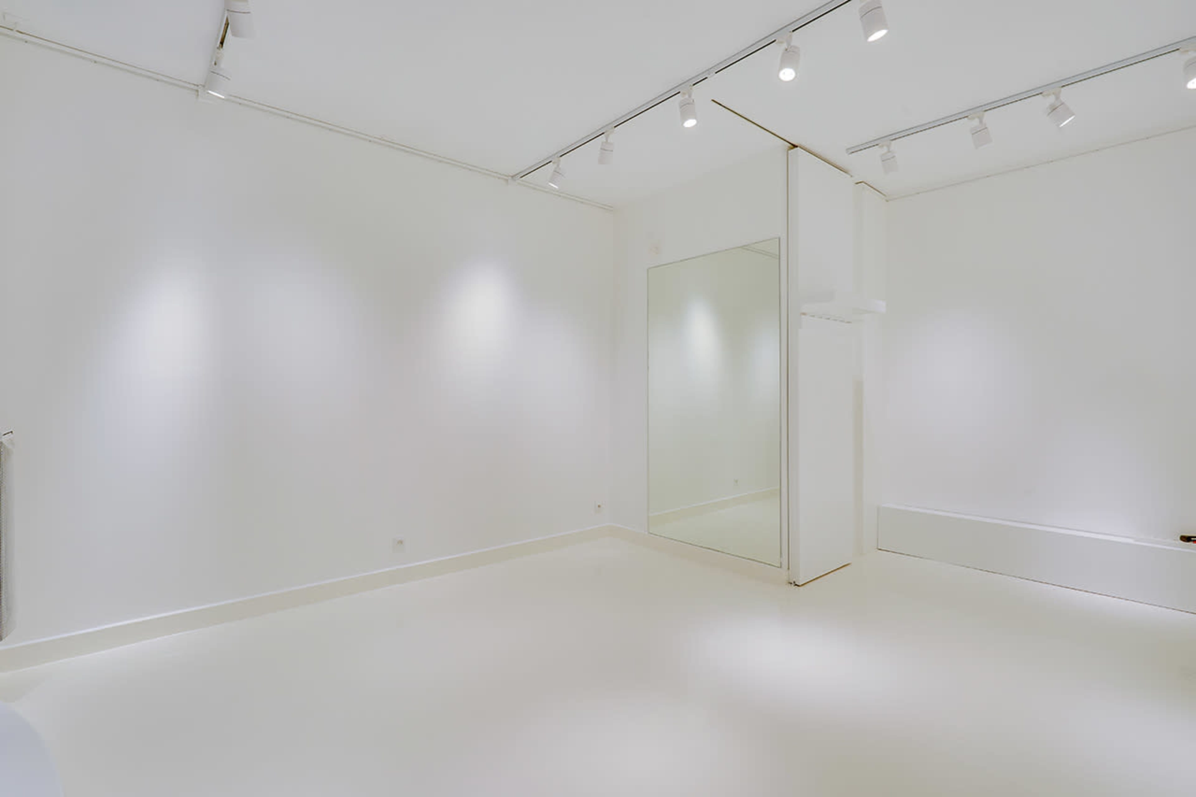The image shows a spacious, minimalist white room with track lighting and a large mirror on the wall.