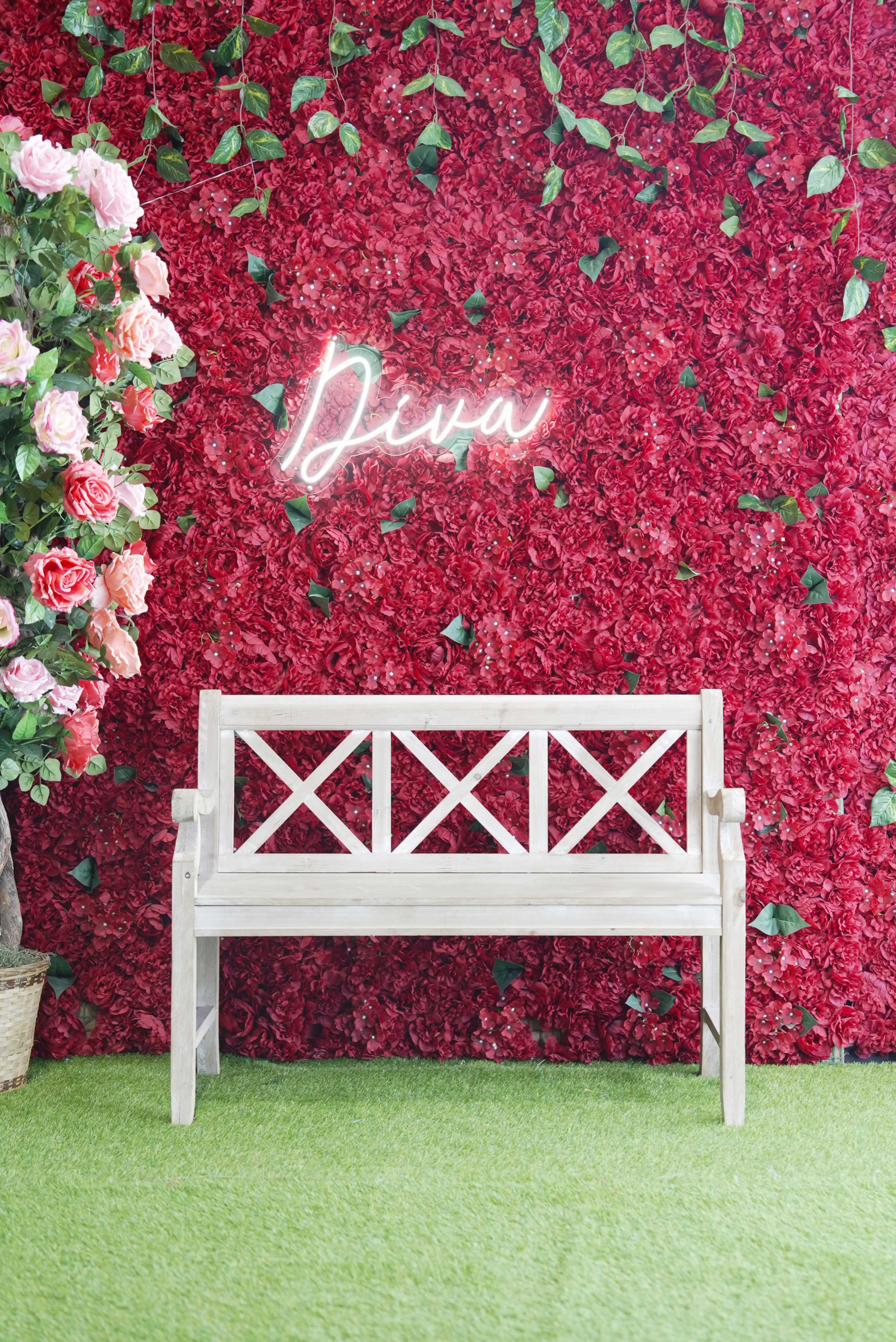 The image shows a white wooden bench in front of a vibrant red flower wall, adorned with a neon sign that reads "Diva."