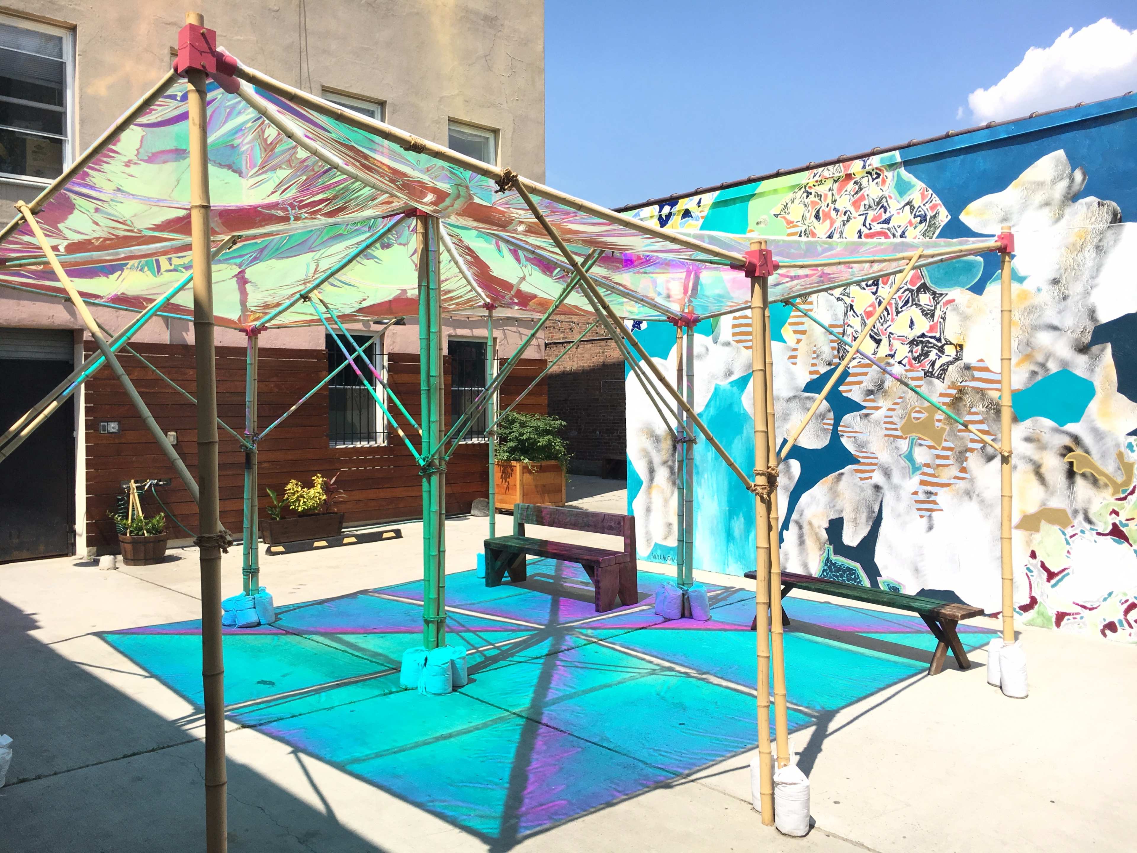 A structure made of bamboo and colorful reflective material stands in an open area with a mural on the wall nearby.