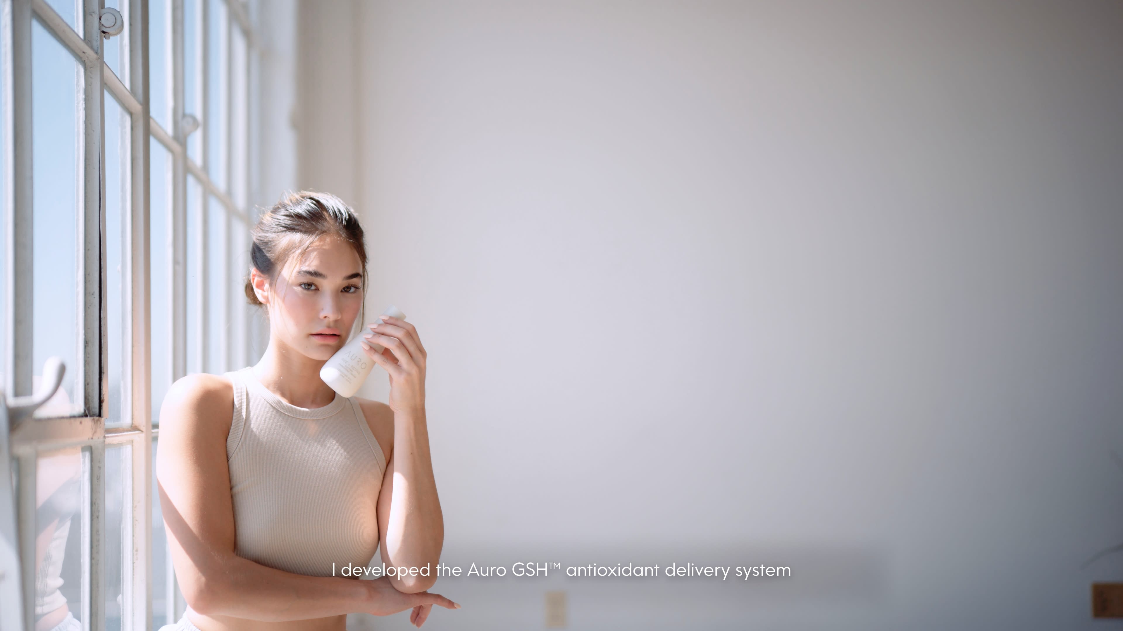 A person stands in a bright, minimalist space while holding a skincare product.