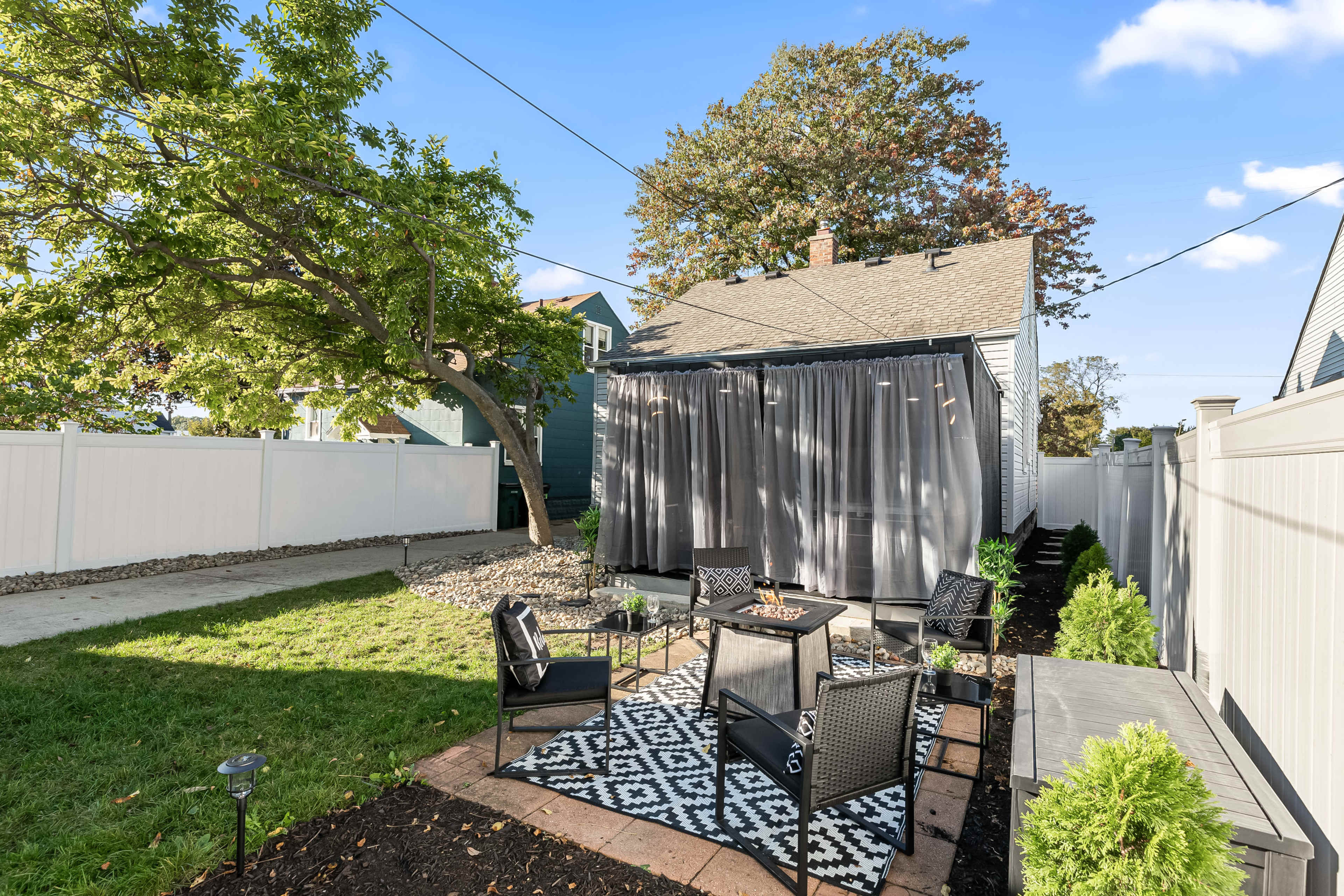 The image shows a backyard area featuring a patio with black furniture, a patterned rug, and a privacy curtain, surrounded by green grass and white fencing.