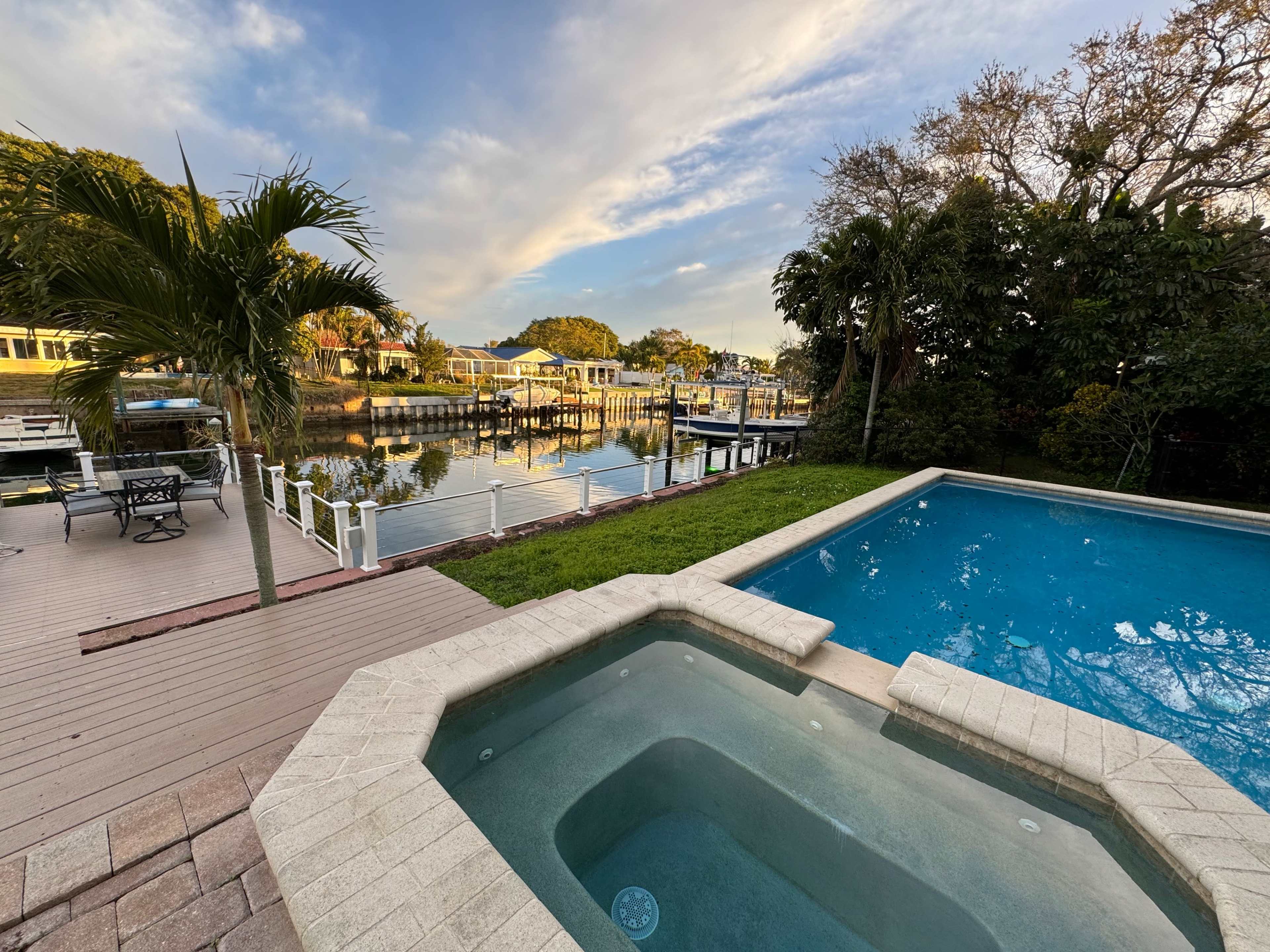 A backyard scene featuring a swimming pool and spa adjacent to a canal with boats and waterfront homes in the background.