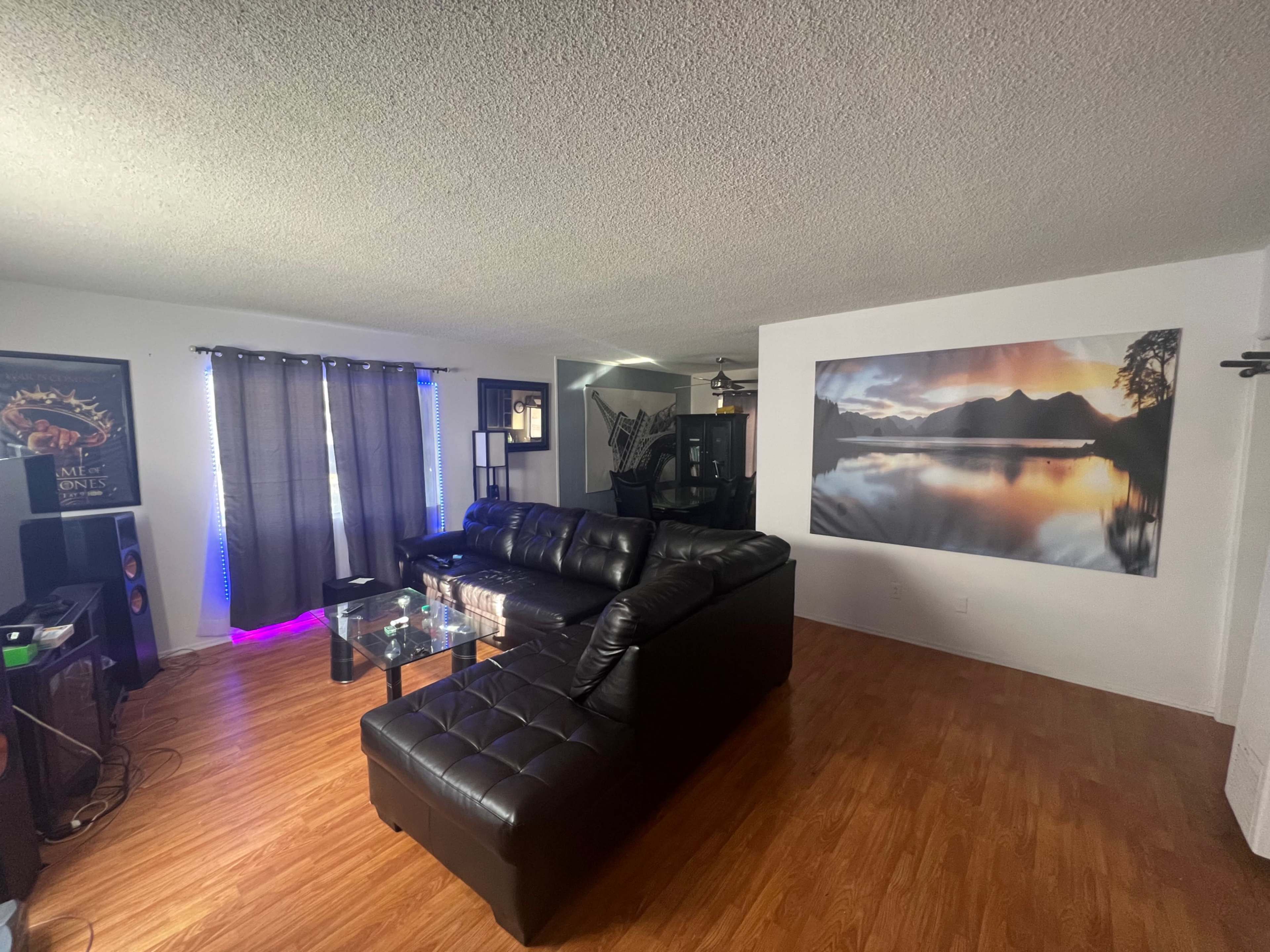 A living room with dark furniture, a large wall-mounted landscape photograph, and a window with partially drawn curtains.