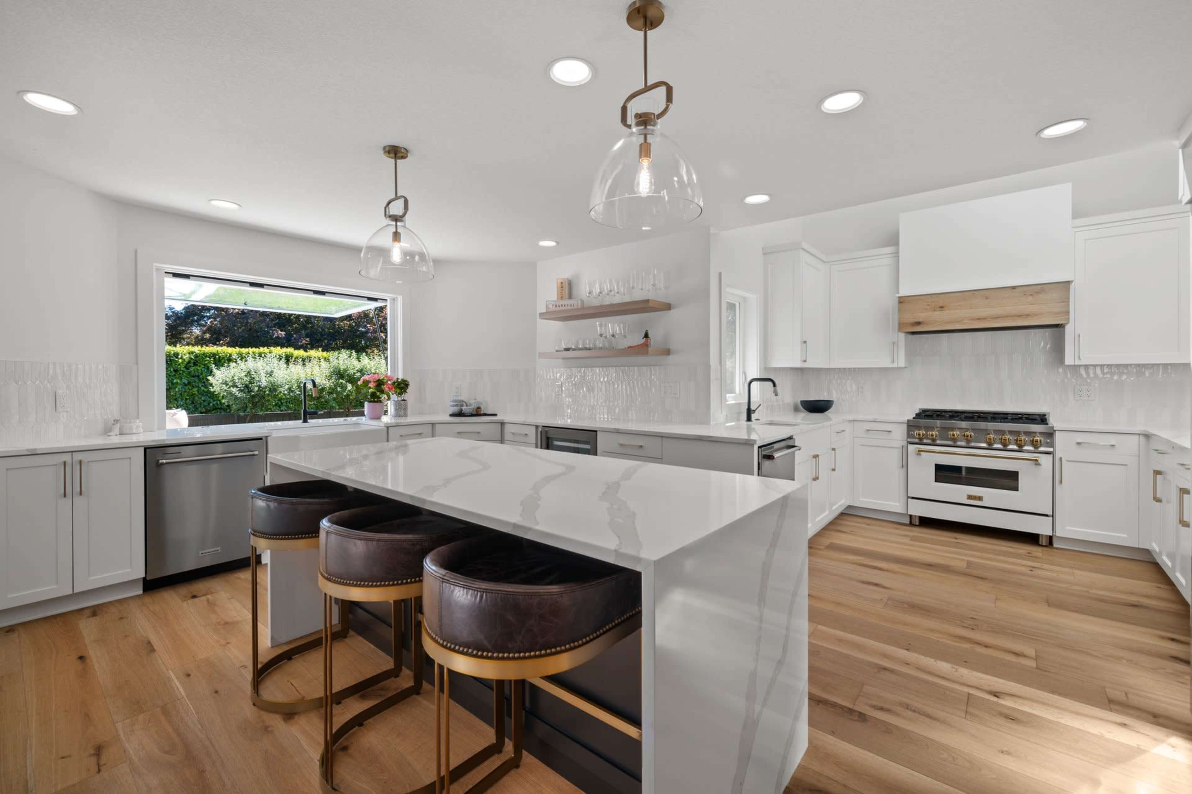 A modern kitchen features a spacious island with three stools, stainless steel appliances, and large windows overlooking a garden.