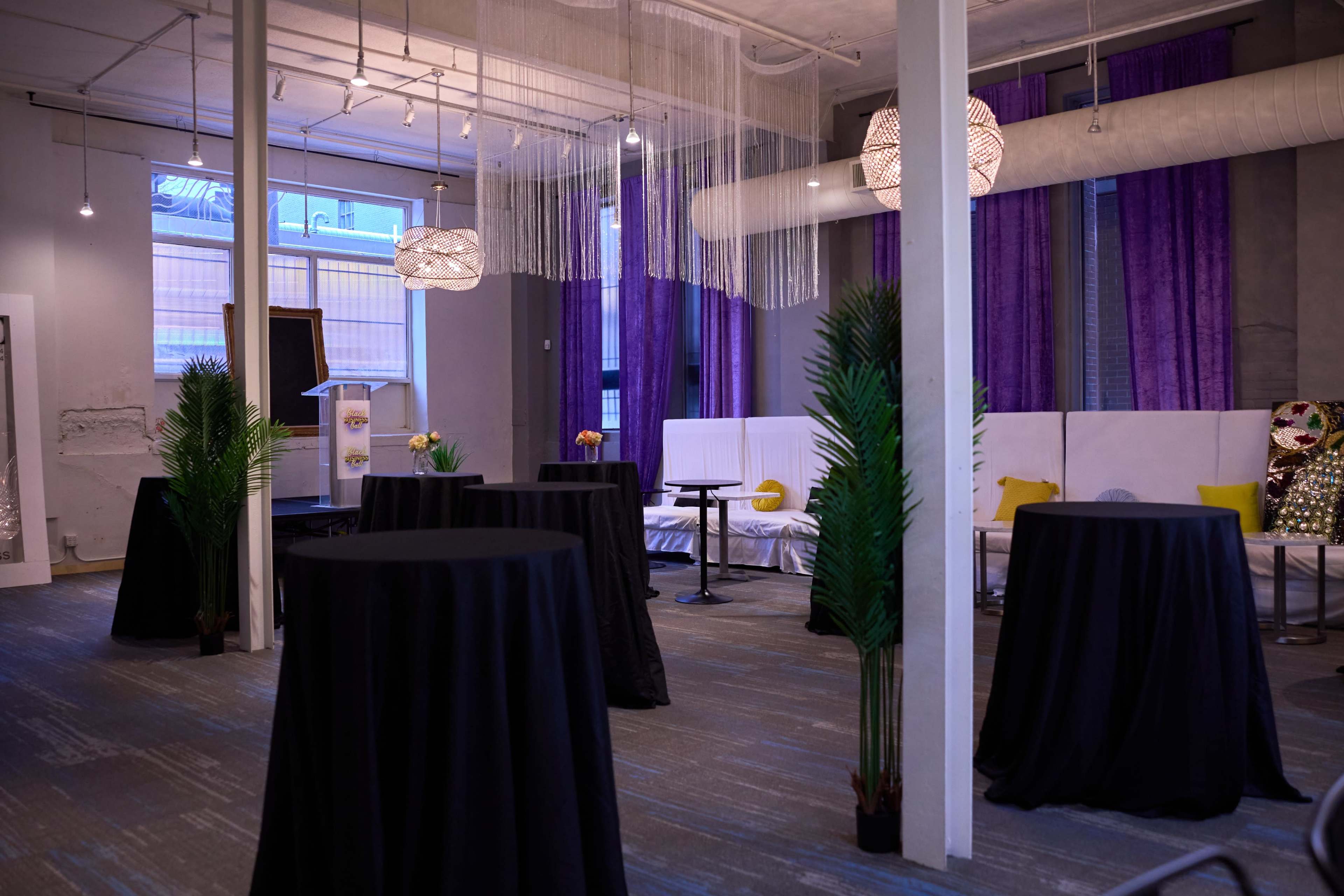 The image shows an elegantly arranged event space with dark round tables, decorative plants, and purple curtains.