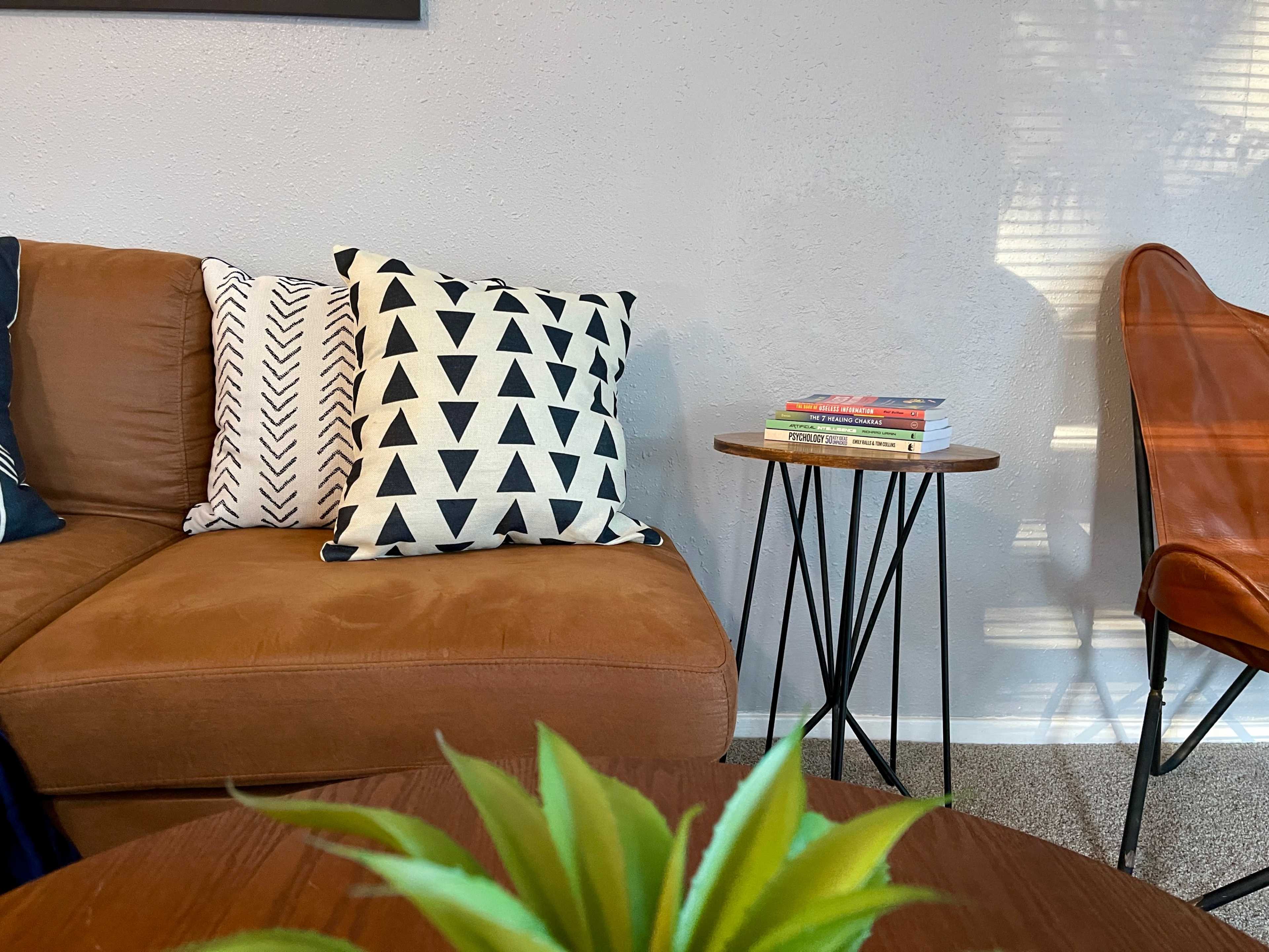 A brown sectional sofa with geometric patterned pillows sits beside a round wooden table holding a stack of books and a small green plant.
