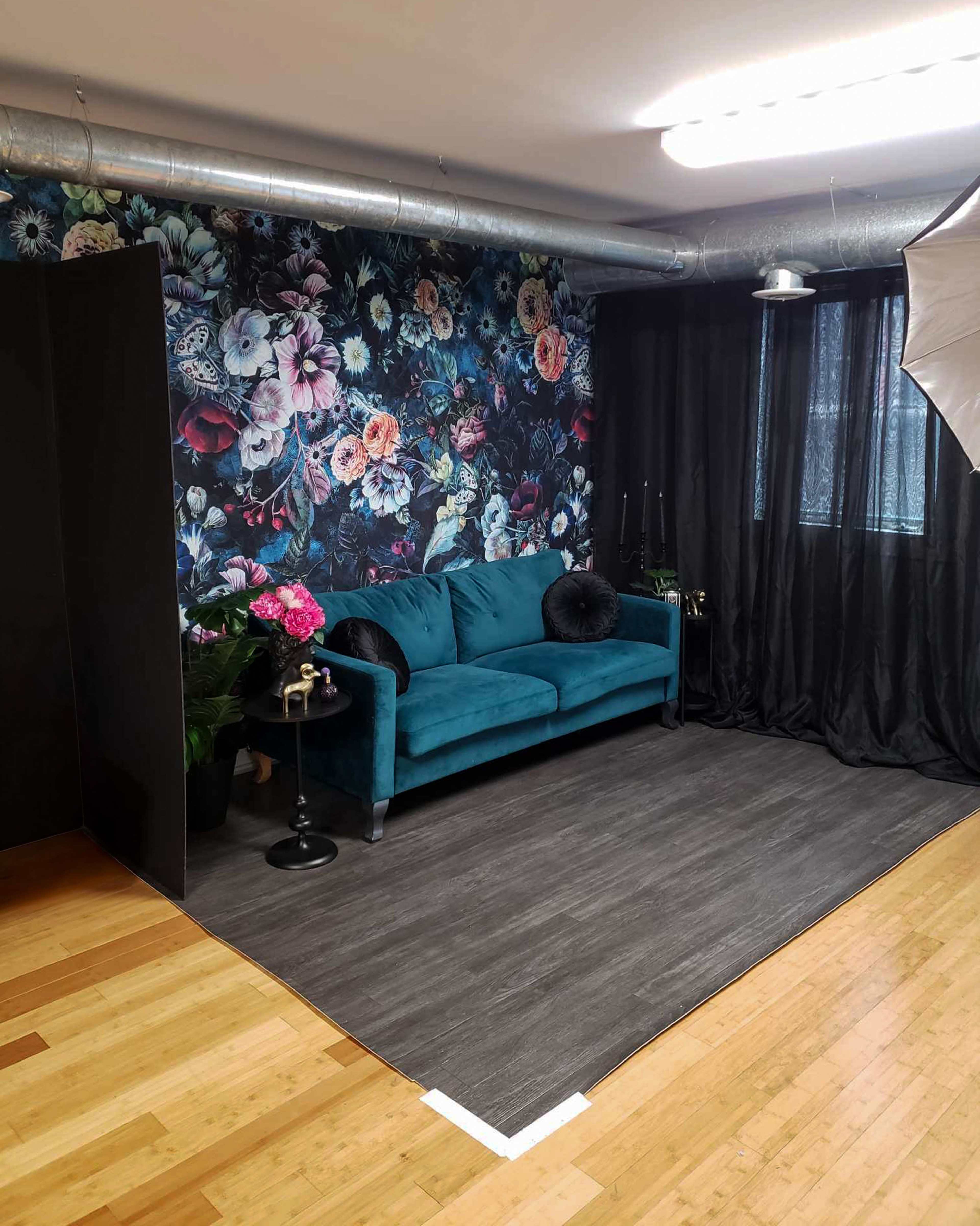 The image shows a photo studio featuring a turquoise couch against a floral wallpaper, with black curtains and hardwood flooring.