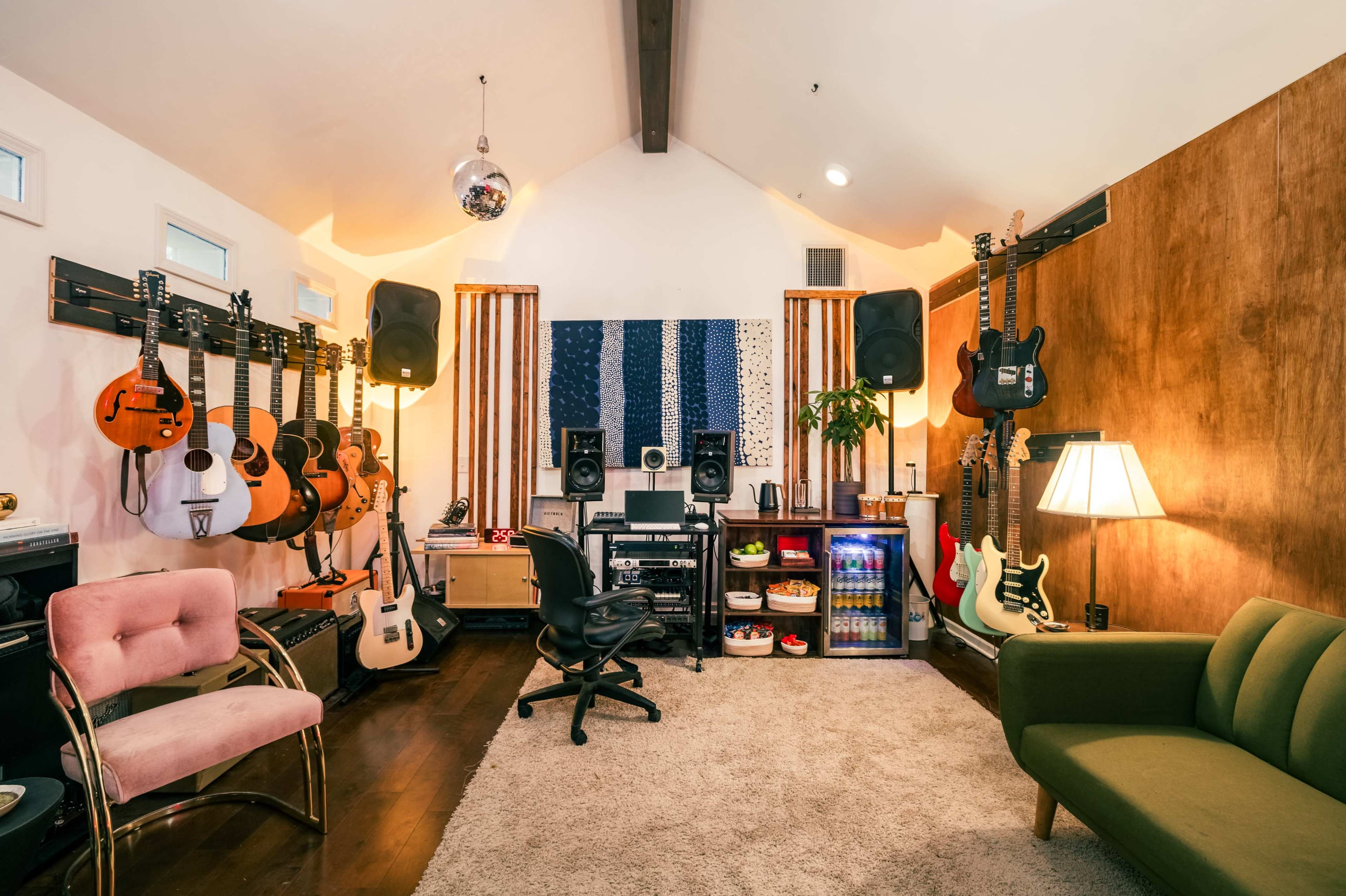 The image shows a music studio with various guitars hanging on the walls, a mixing area with speakers, and a cozy seating arrangement.
