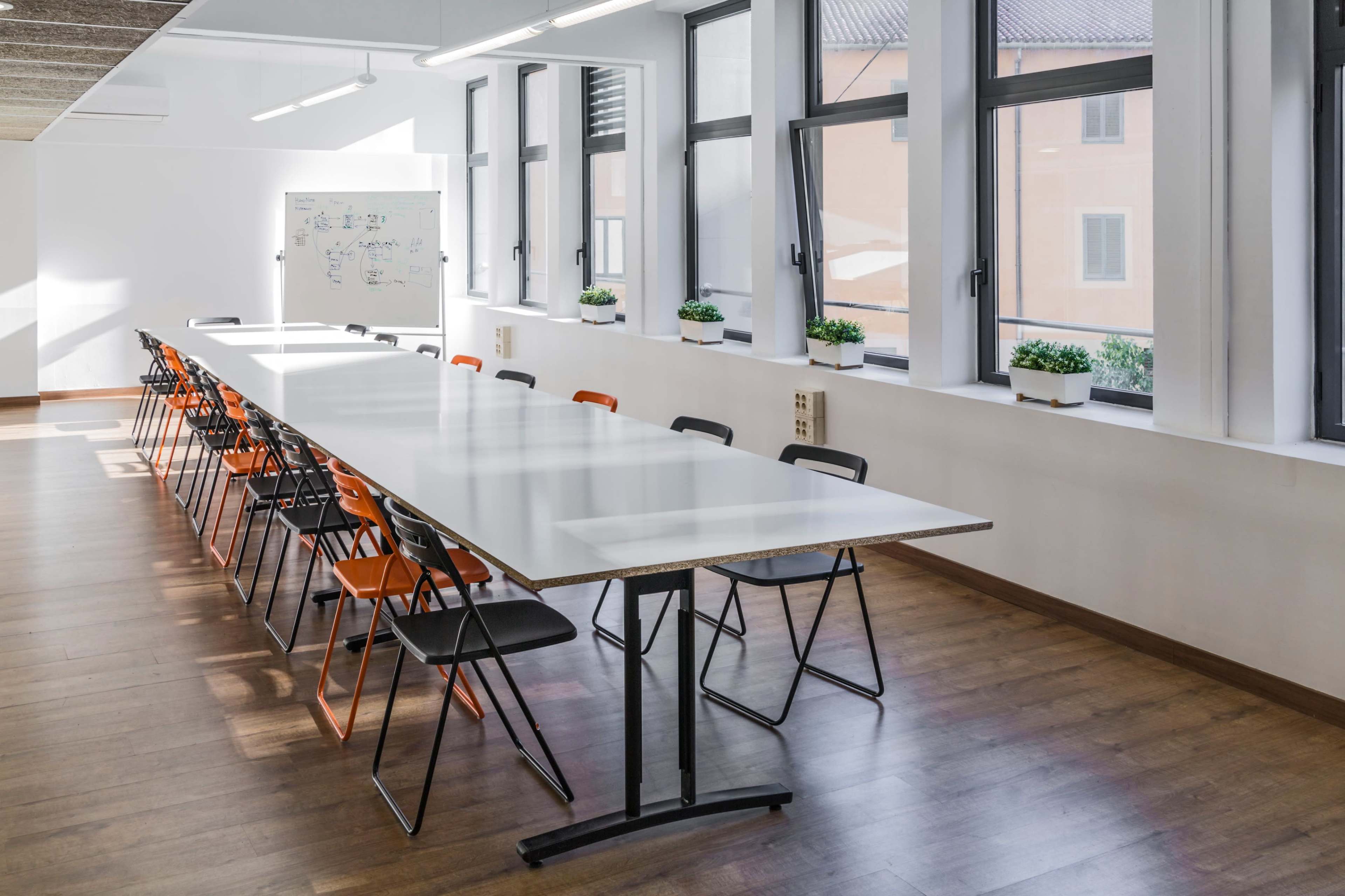 A long, rectangular table with black and orange chairs is set up in a well-lit meeting room featuring large windows and a whiteboard.