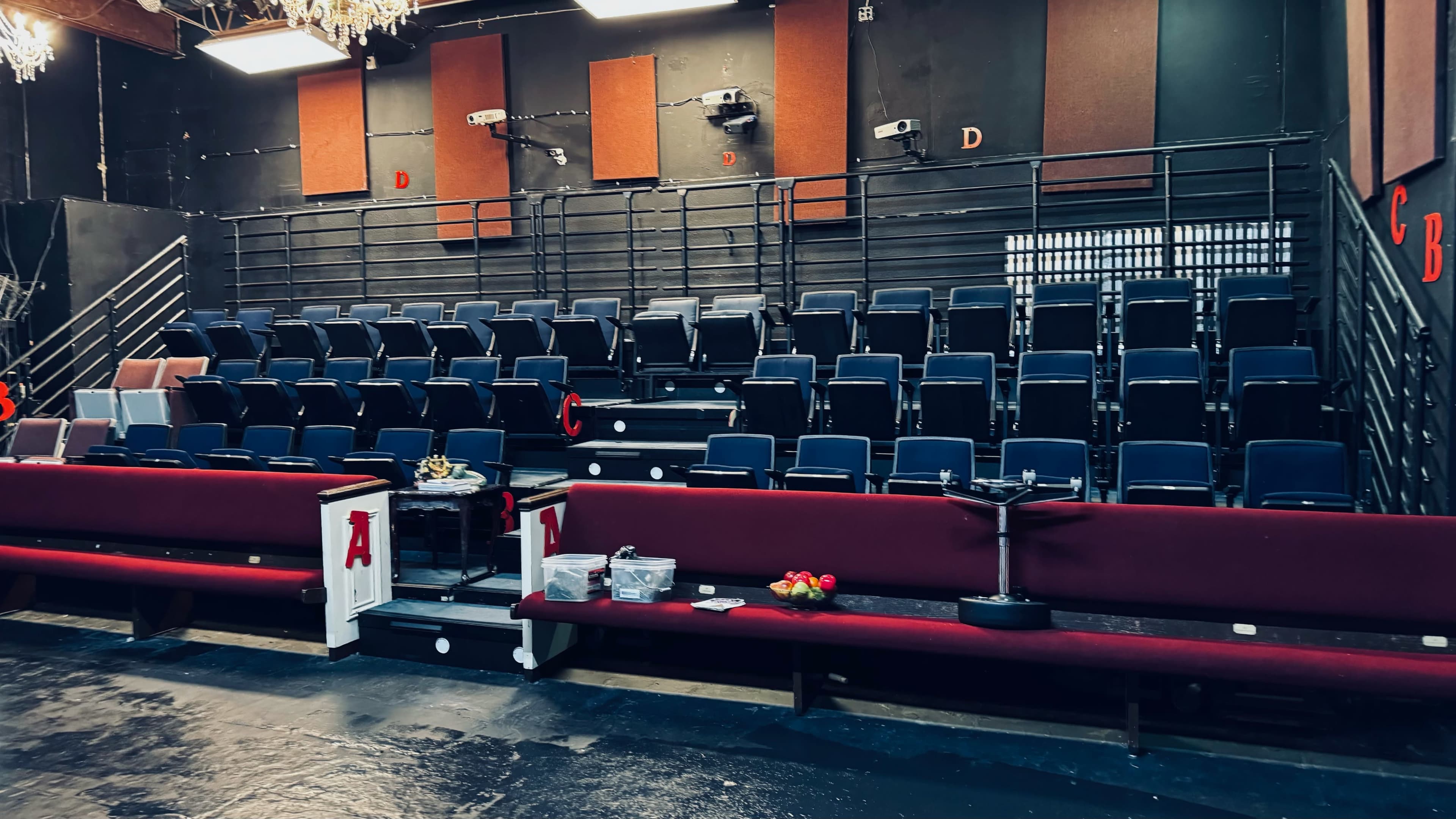 A seating area in a theater features dark blue chairs arranged in rows with maroon benches in front, along with some boxes and items on the ground.