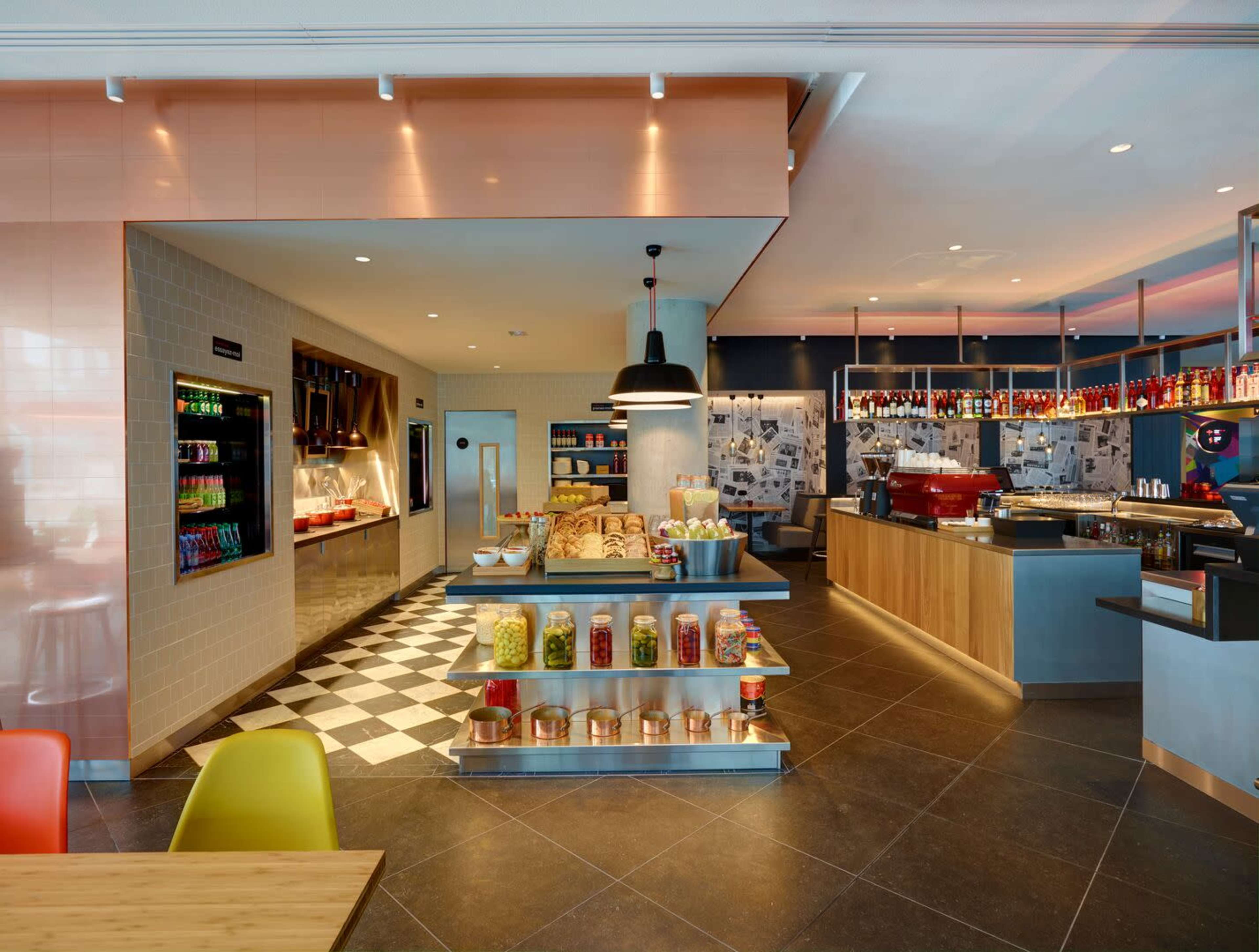 A modern café interior with a display counter filled with various baked goods, a beverage station, and seating areas featuring colorful chairs.