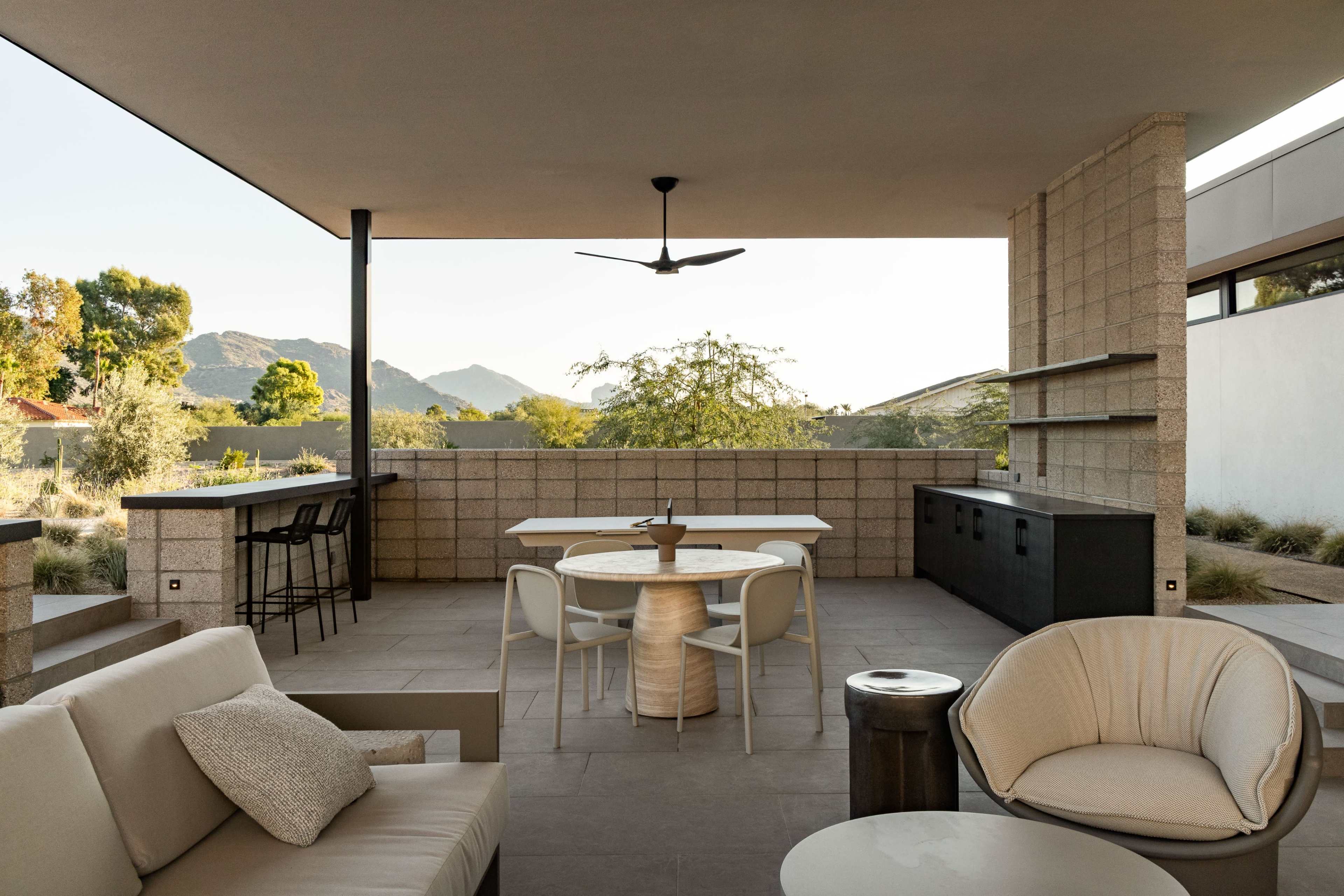 The image shows a modern outdoor patio with a dining area and seating, surrounded by mountains in the background.