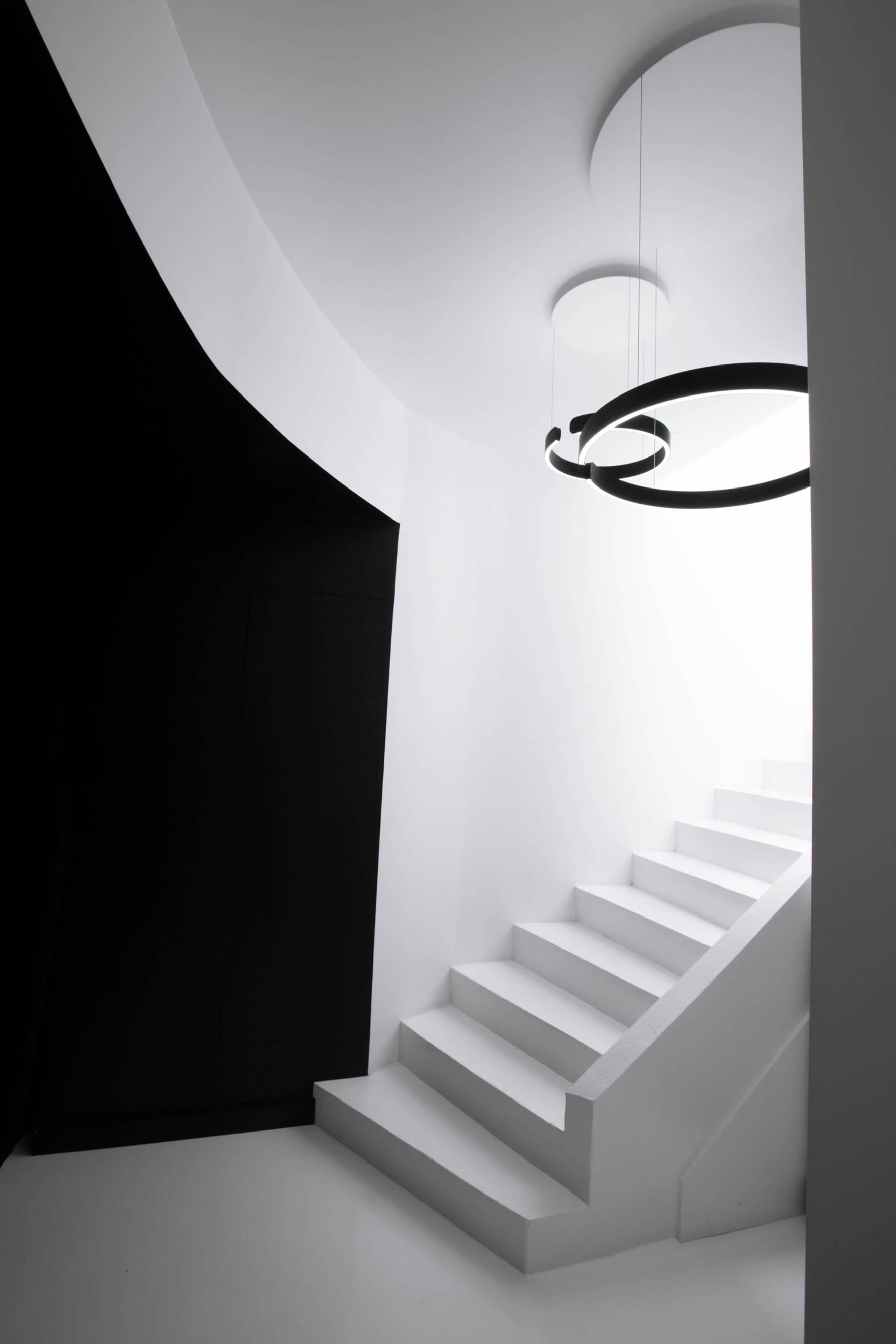 A minimalist staircase ascends in a bright white space, illuminated by a circular pendant light against a contrasting black wall.