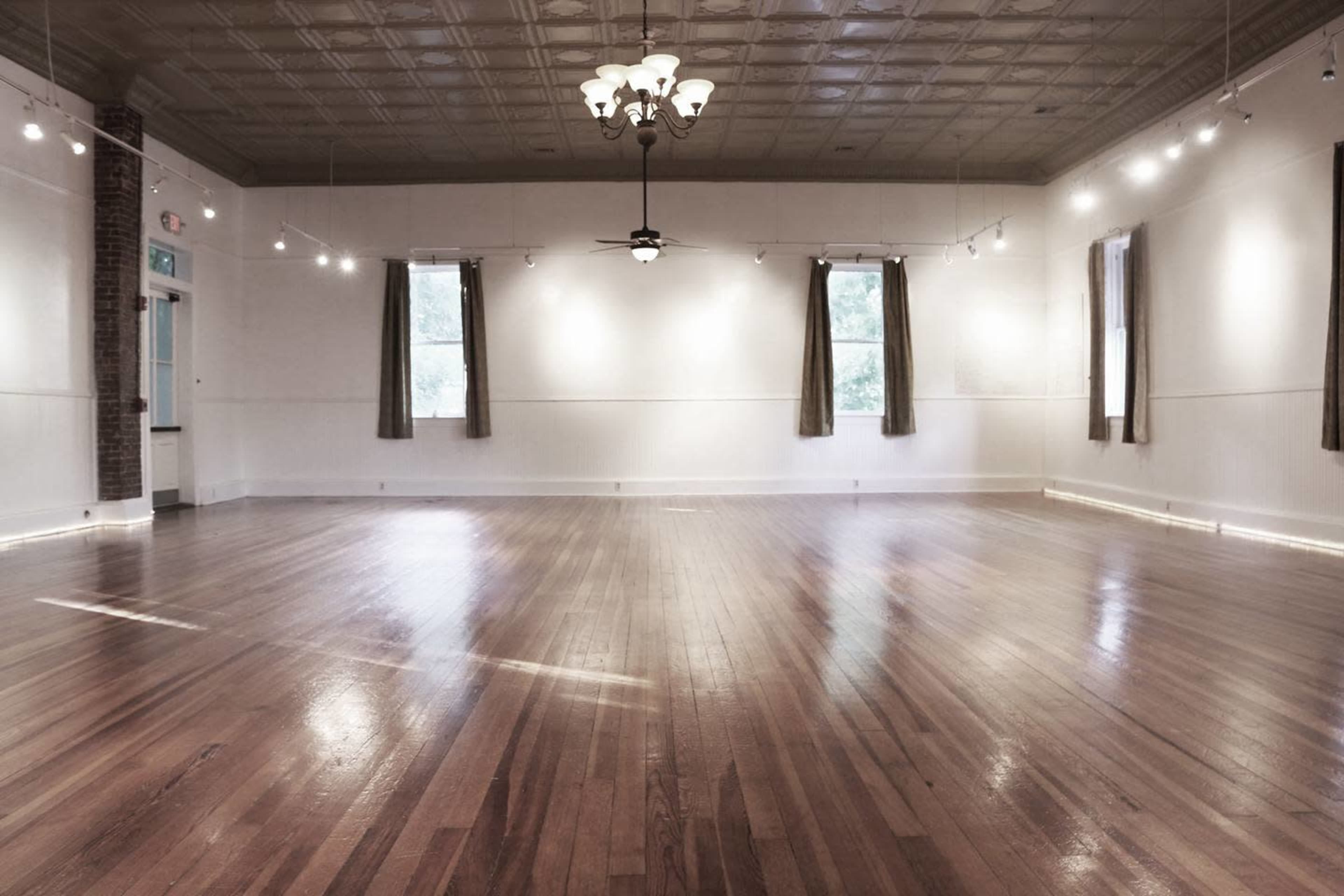 The image shows a spacious, well-lit interior room with wooden floors and large windows featuring curtains.