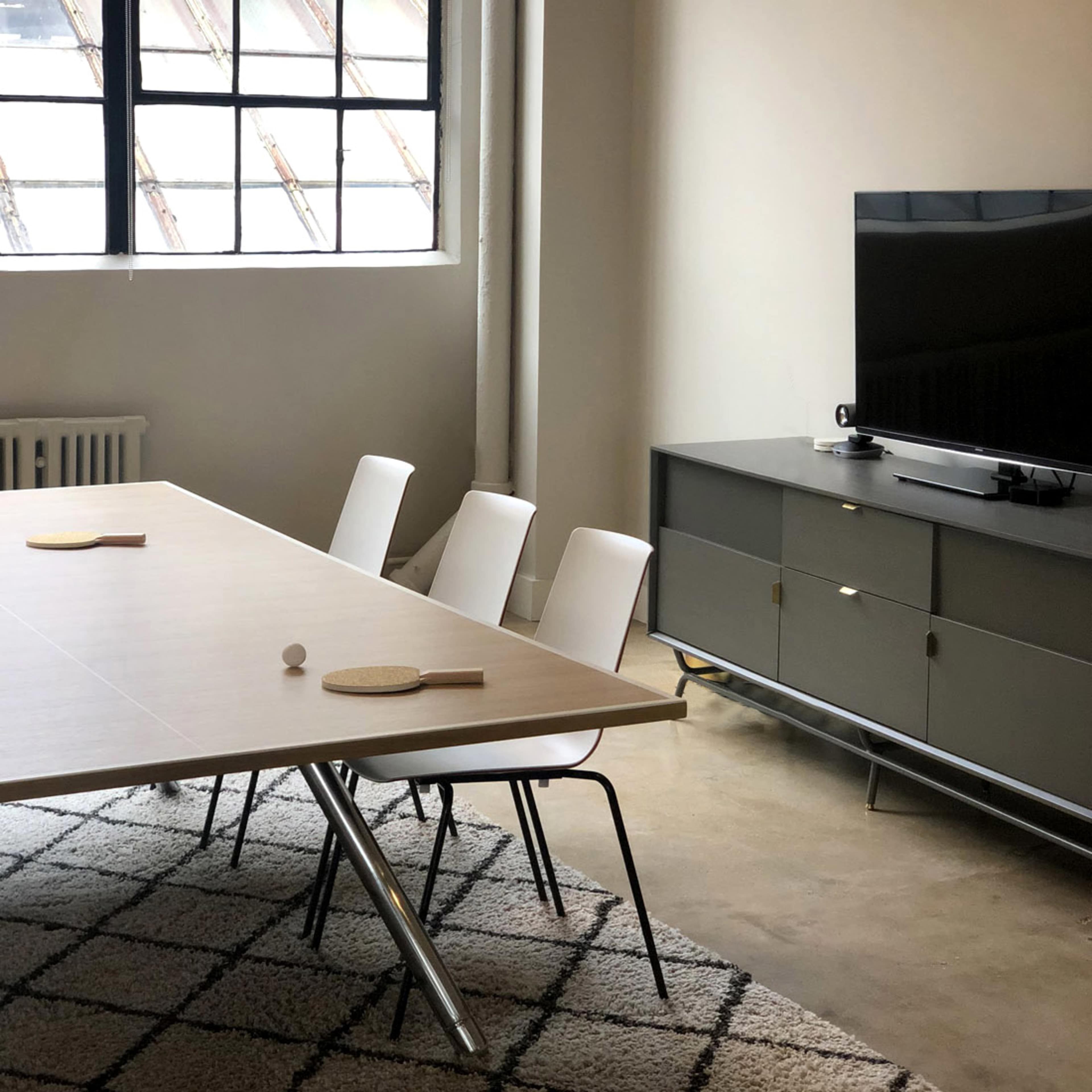 A minimalist room features a large wooden table with four white chairs, a ping pong paddle, a ball on the table, and a television against a gray cabinet.