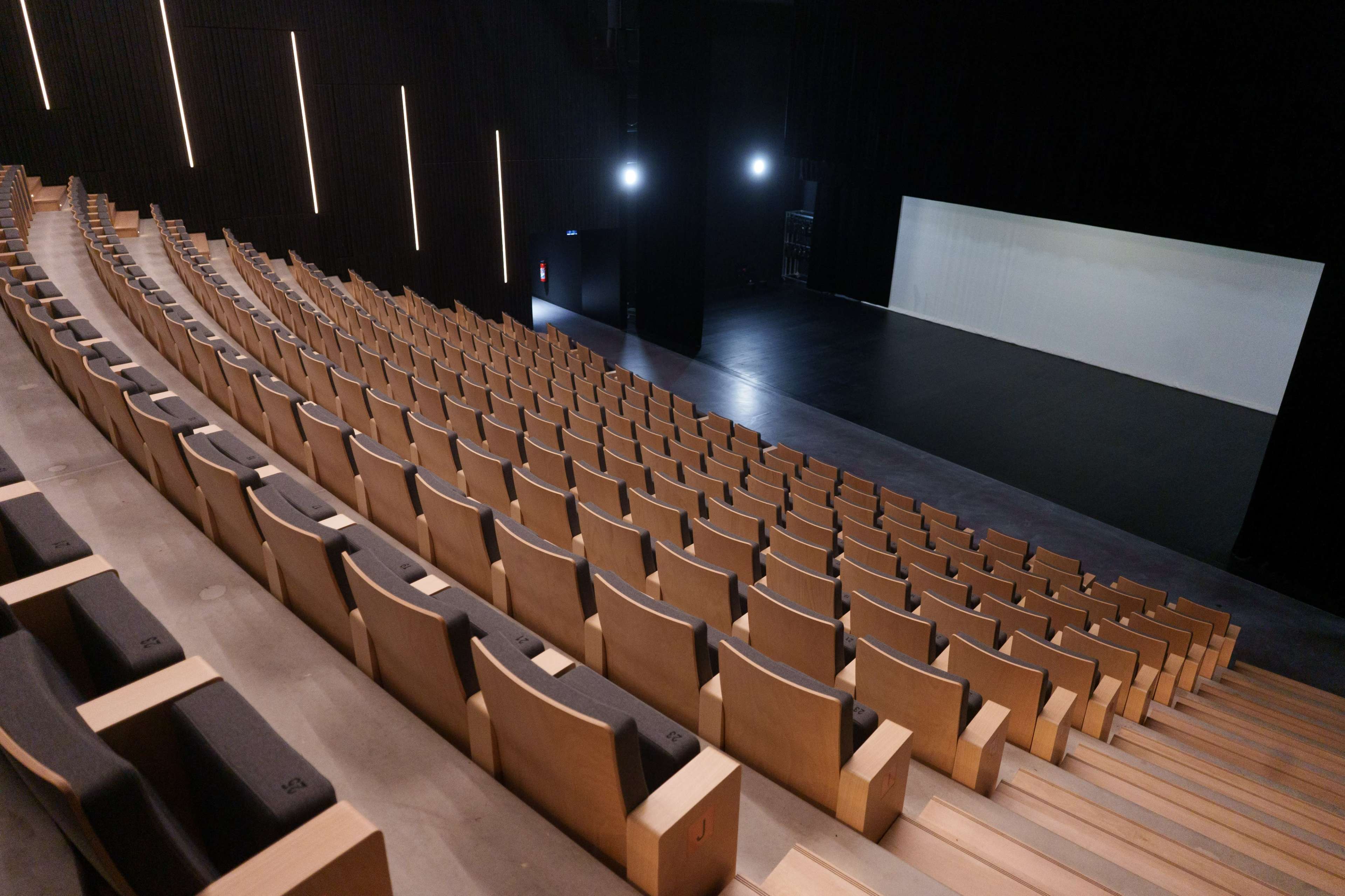 A modern auditorium with tiered wooden seating facing a spacious stage area illuminated by focused lighting.
