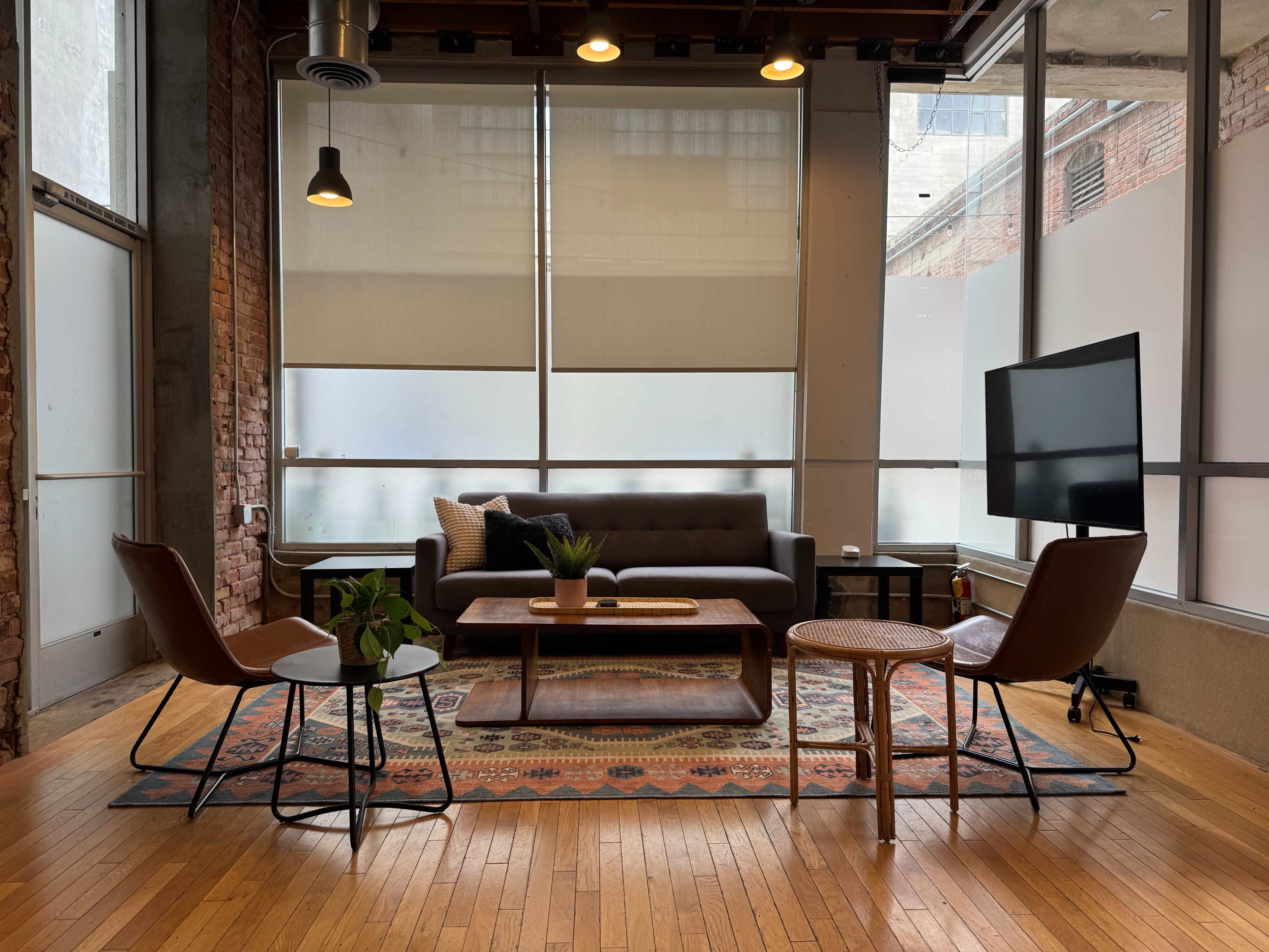 A modern living area features a gray sofa, a wooden coffee table, and four chairs arranged around a patterned rug, with large windows providing natural light.