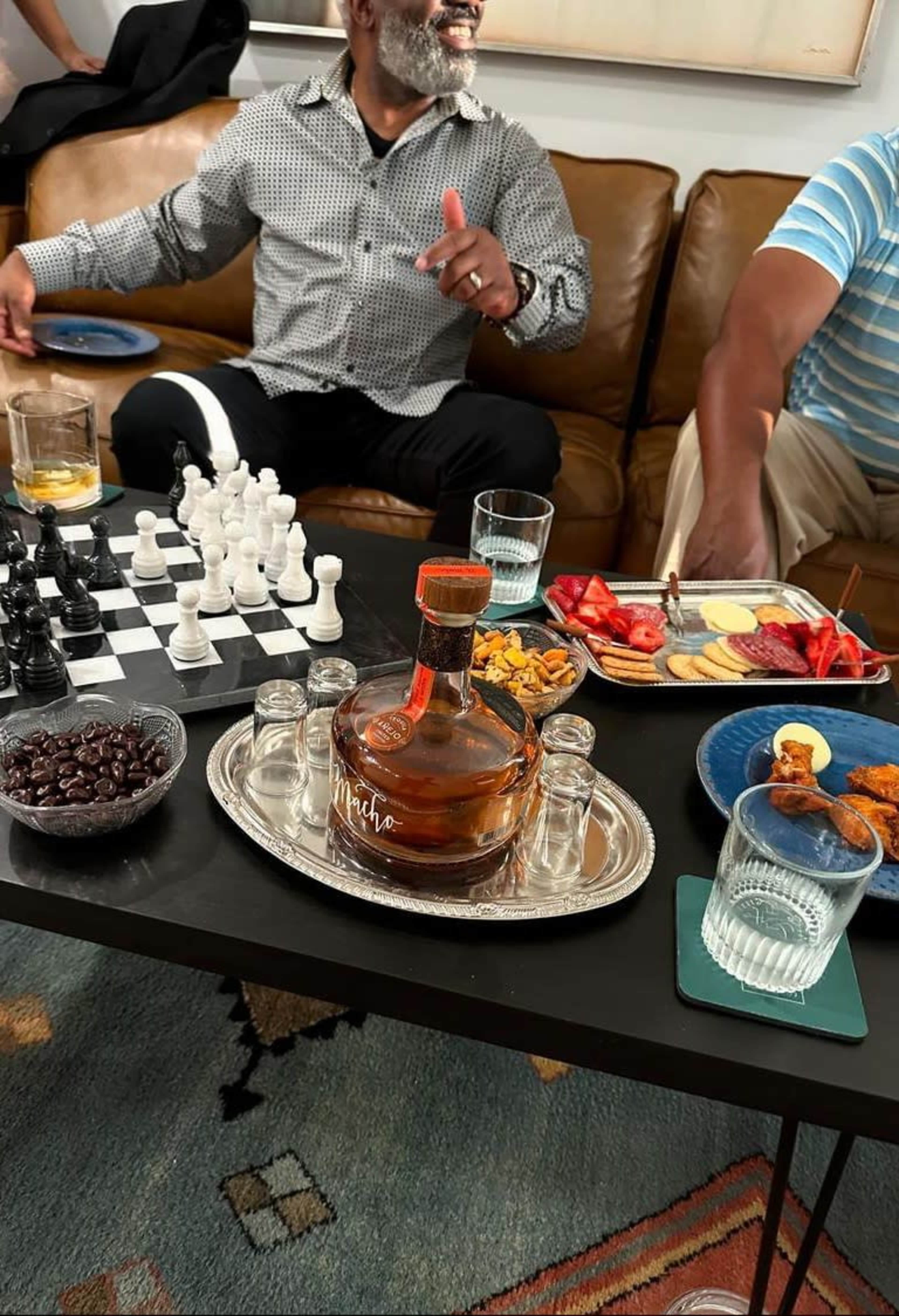 Two men are seated on a sofa with a chessboard and various snacks displayed on a coffee table, alongside drinks and a bottle of liquor.