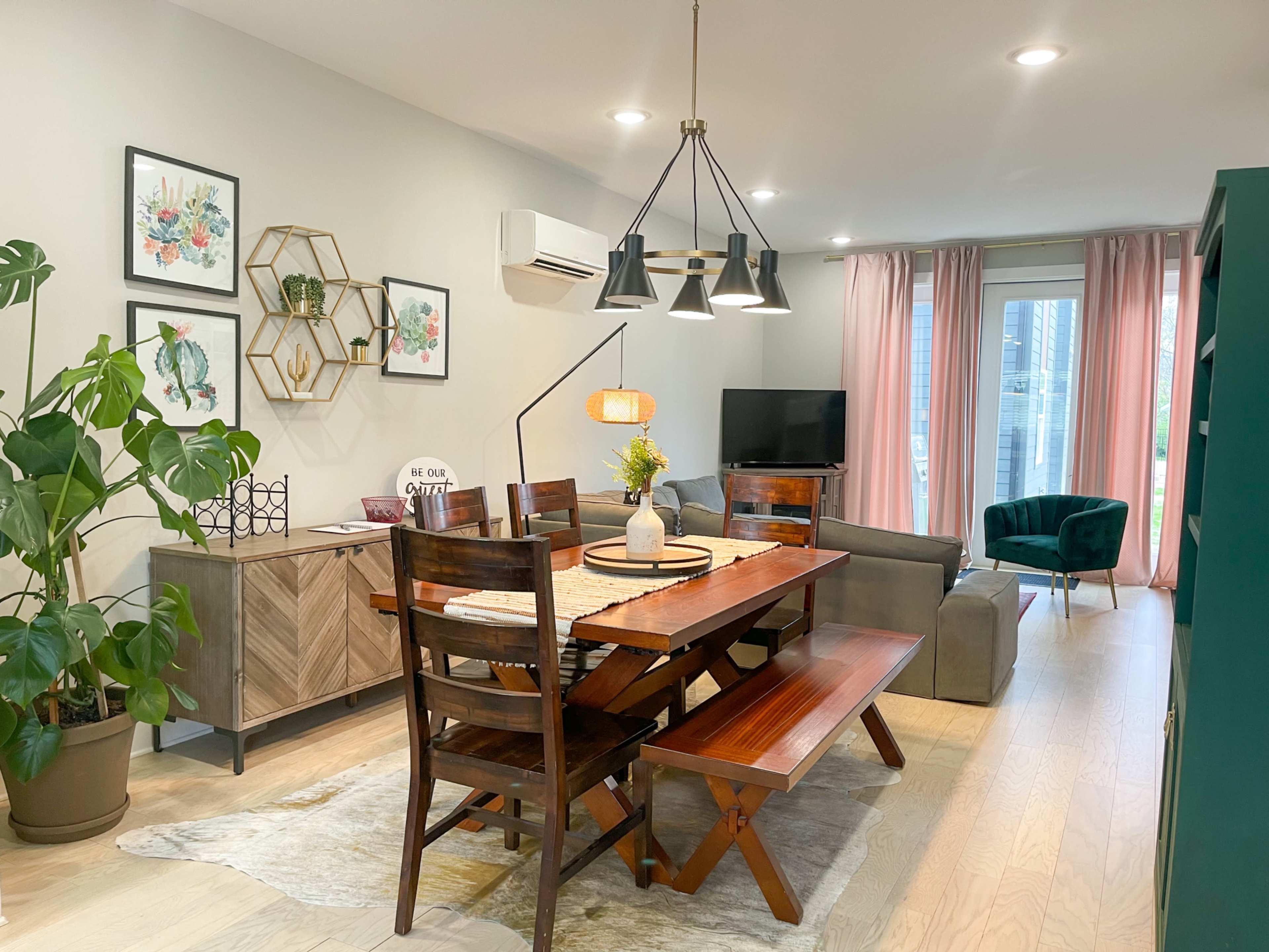 The image shows a dining area with a wooden table and benches, surrounded by decorative plants, artwork on the walls, and a living area with a couch and television in the background.