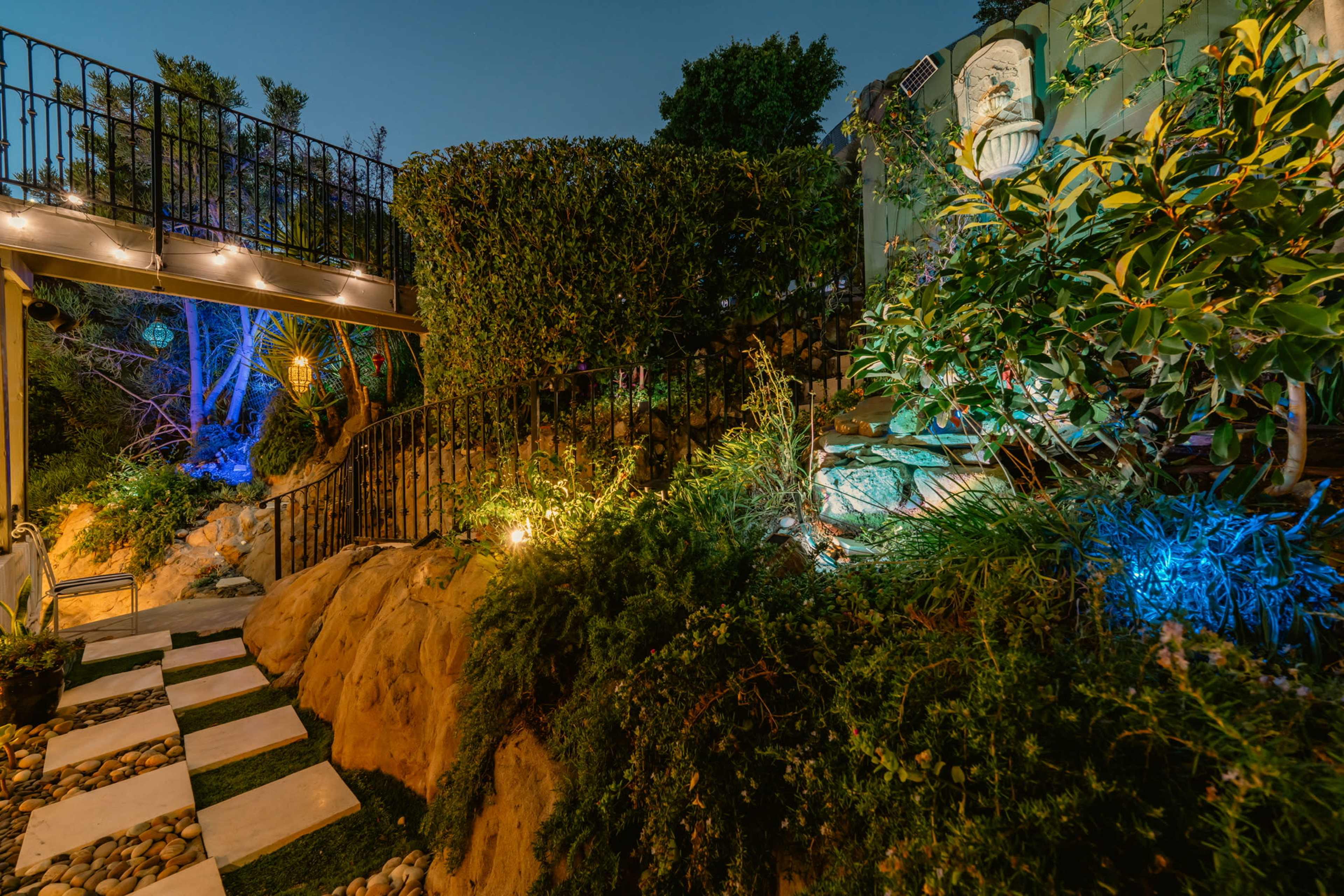 A landscaped pathway with stepping stones winds through greenery, leading to a stone wall adorned with a statue, illuminated by soft outdoor lighting.