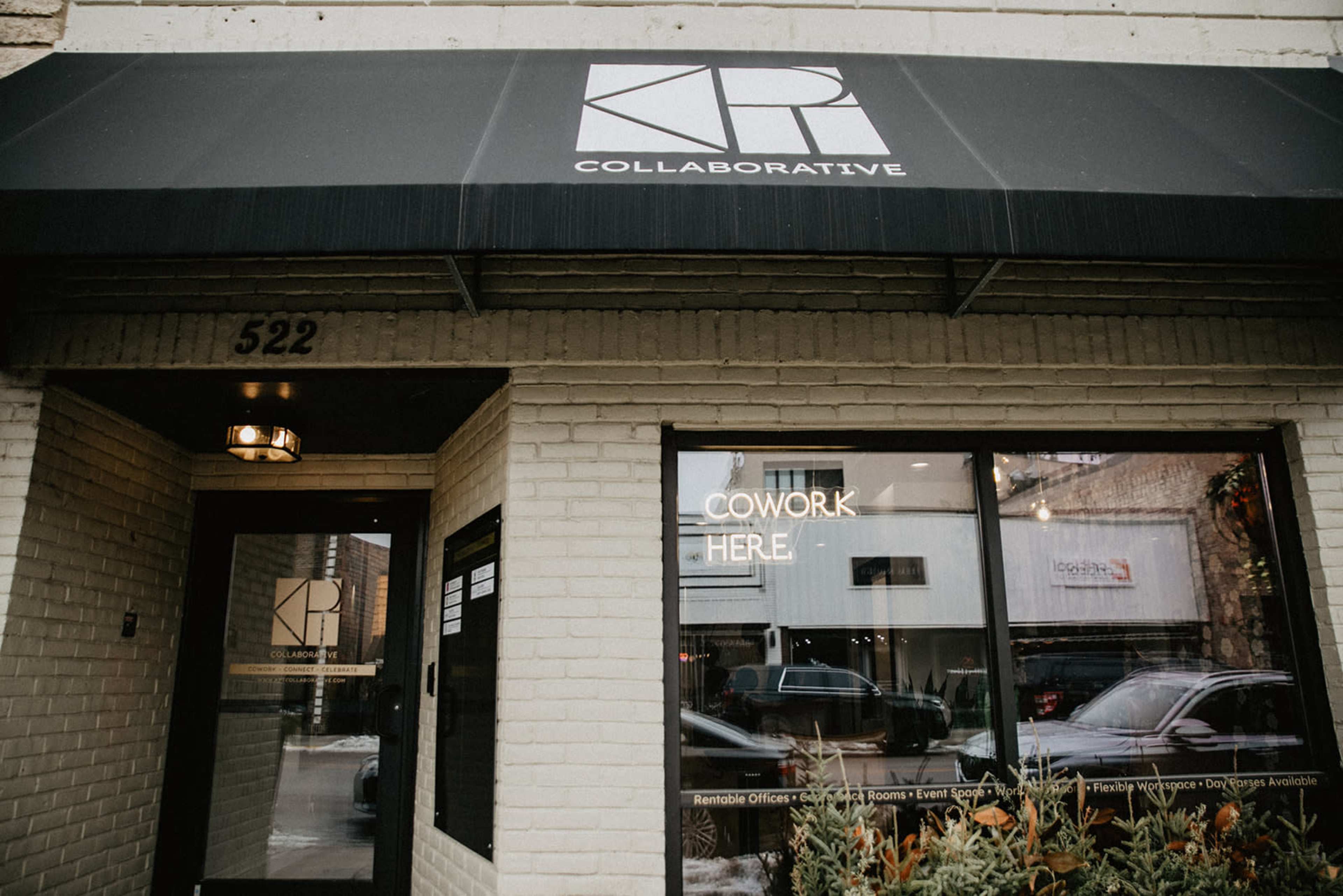 The image shows the entrance of KPI Collaborative, a coworking space, with a black awning and large glass windows displaying the words "COWORK HERE."