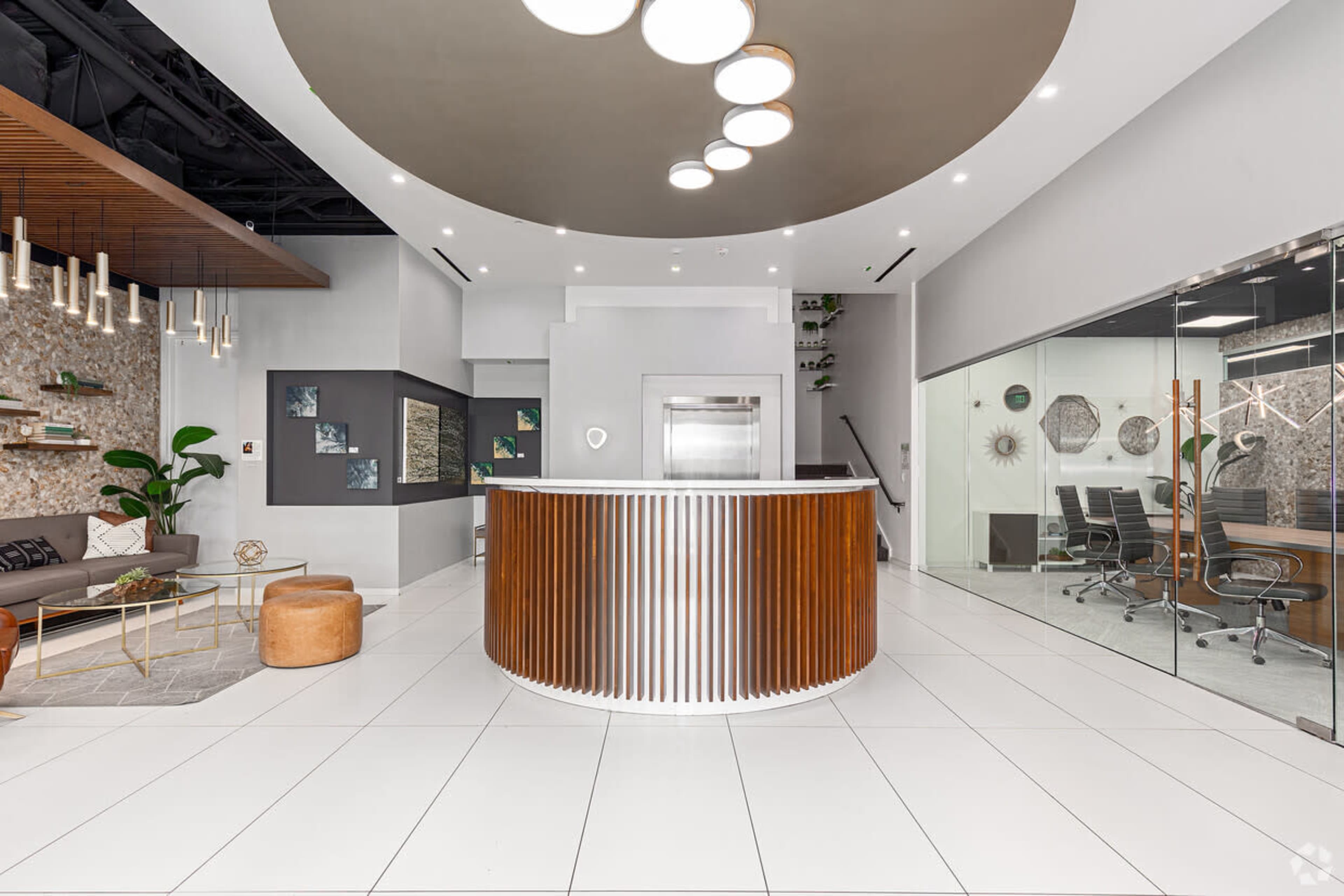 The image shows a modern reception area with a round wooden front desk, white tiled floors, and a glass-walled conference room visible in the background.