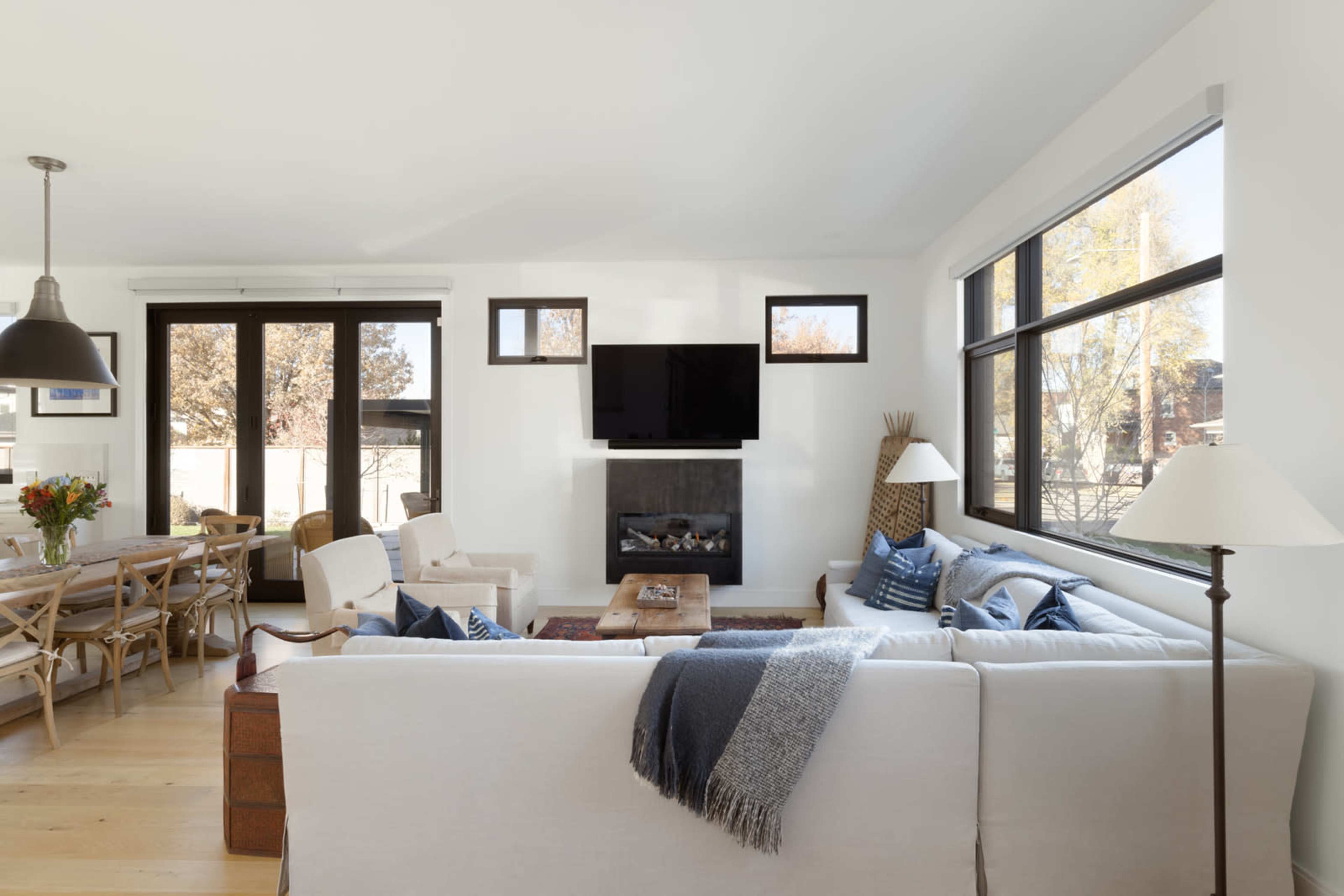 A modern living room features a white sofa, a television mounted on the wall, a fireplace, and large windows letting in natural light.