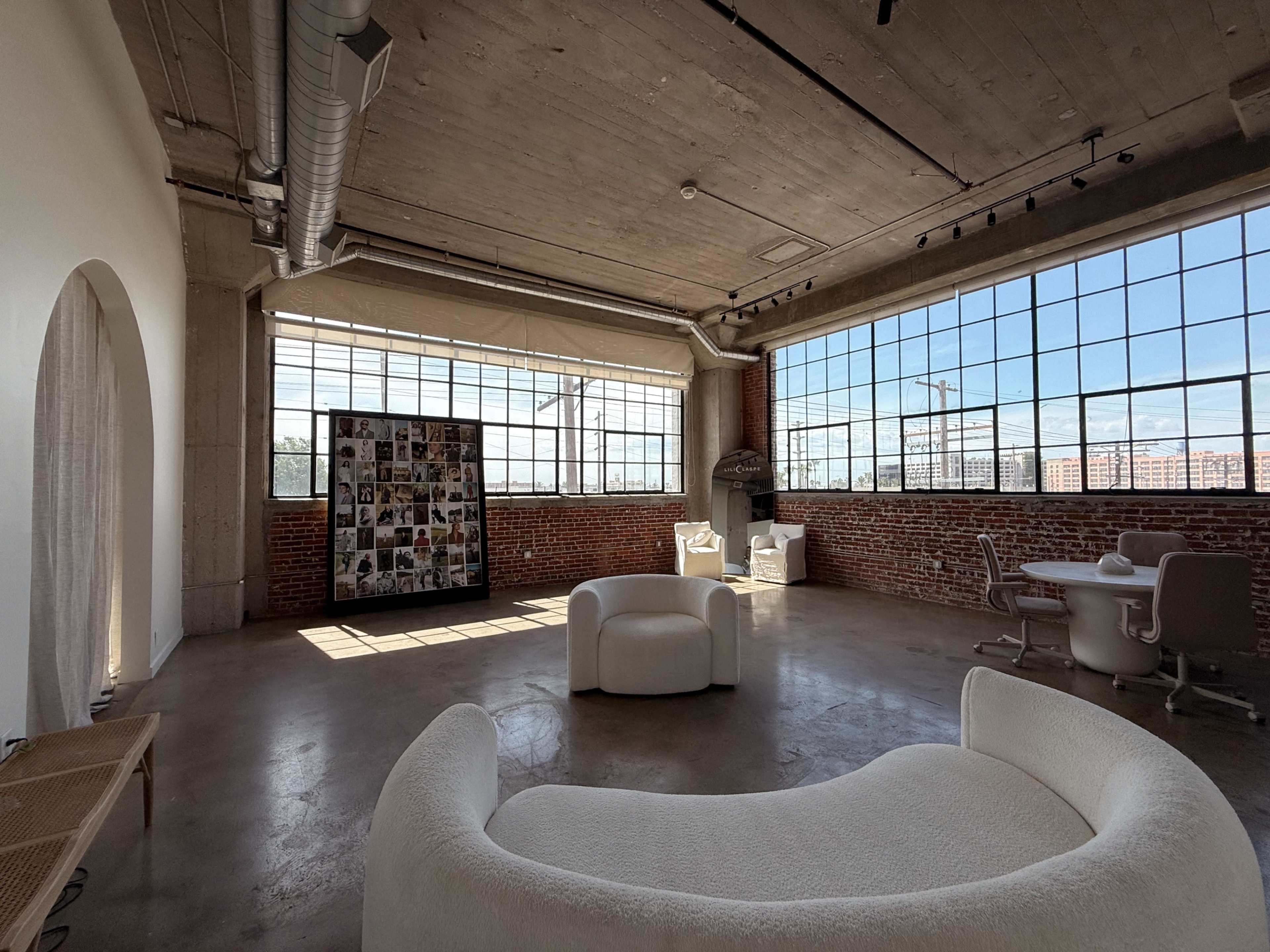 The image shows a spacious, industrial-style room with large windows, brick walls, and contemporary furniture arranged around a central photo display board.