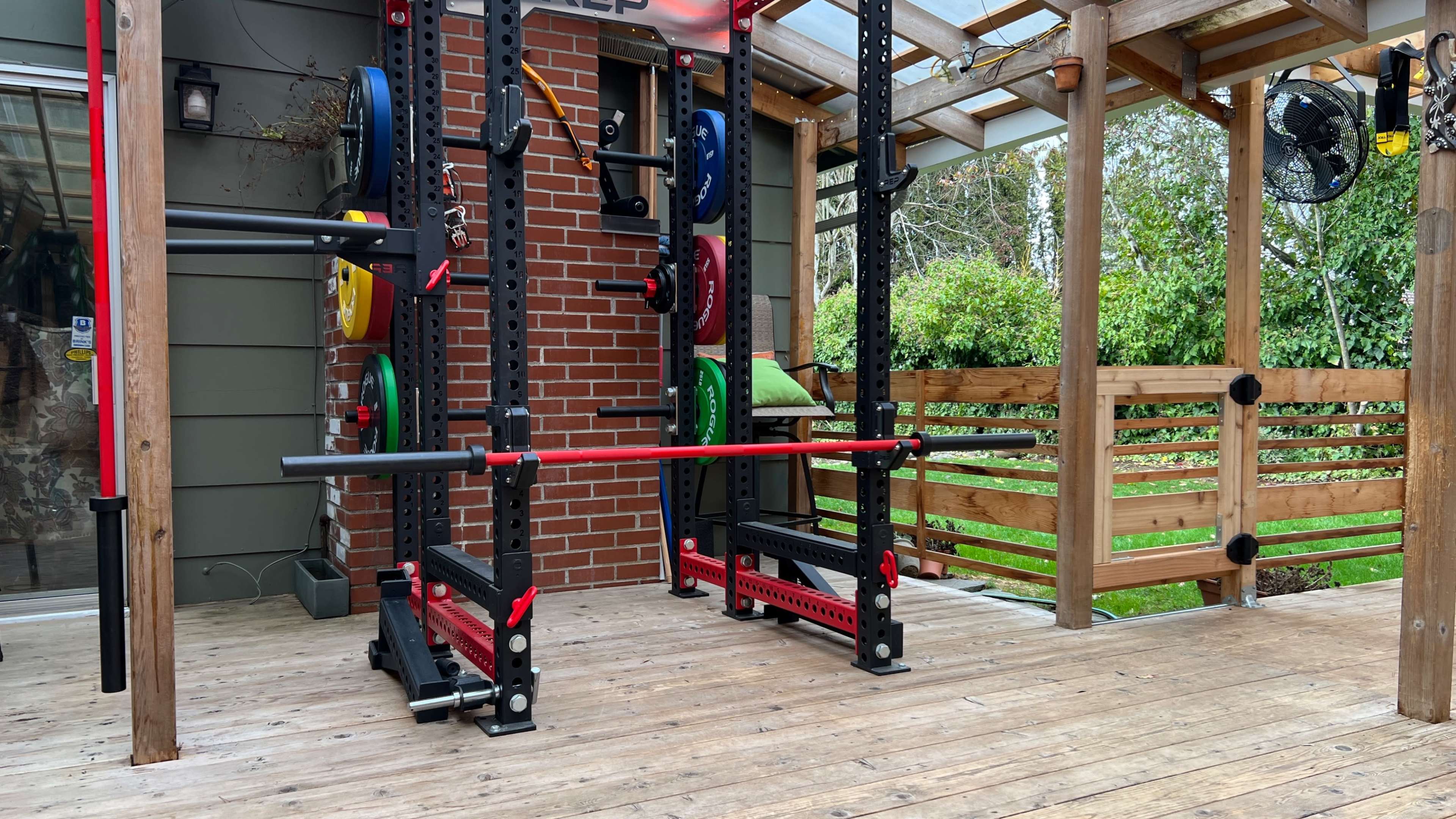 A weightlifting rack with various colored plates is set up on a wooden patio beside a brick wall and greenery.