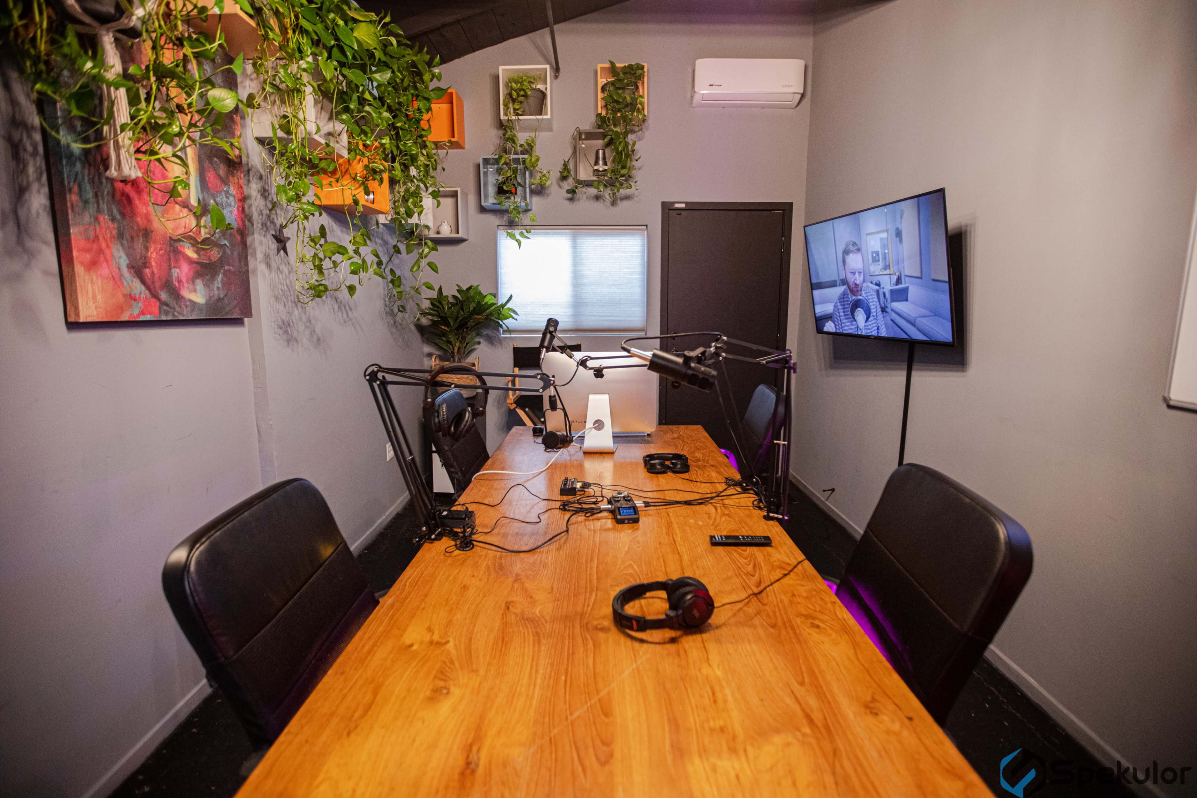 A podcast recording studio features a wooden table with microphone setups, headphones, and wall-mounted greenery.