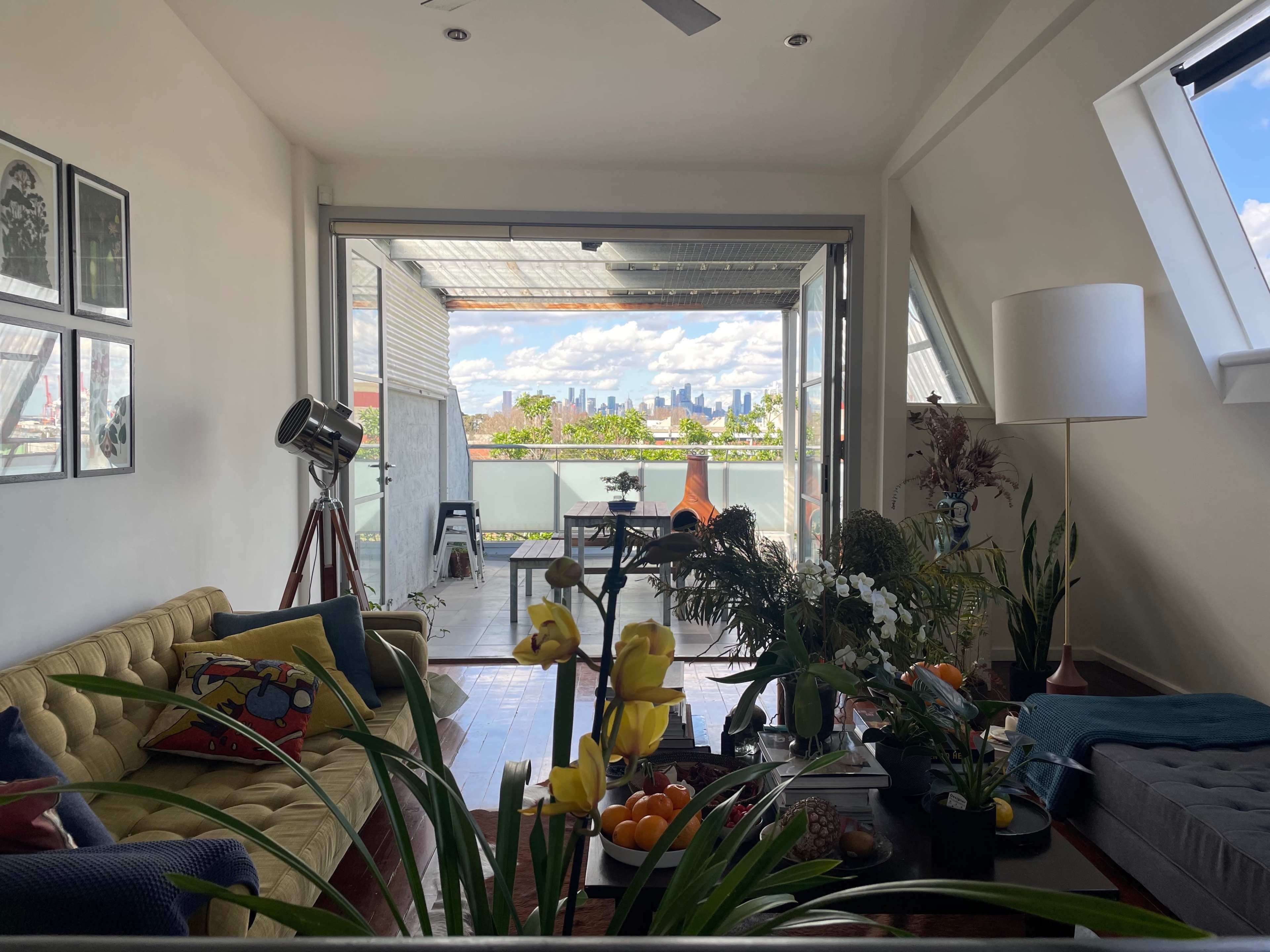 The image shows a modern, open living space with a yellow couch and plants, leading to a balcony that overlooks a city skyline.