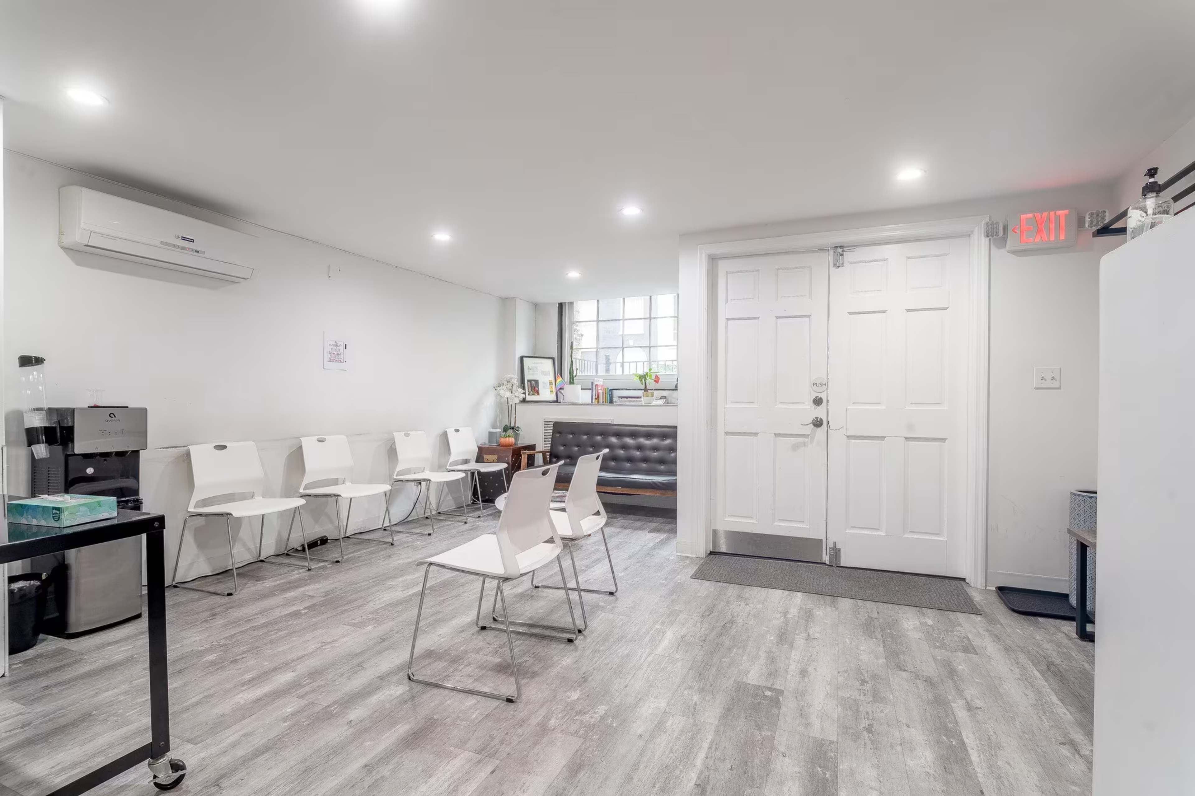 The image shows a modern, well-lit waiting area with white chairs, a reception desk, and an exit door.