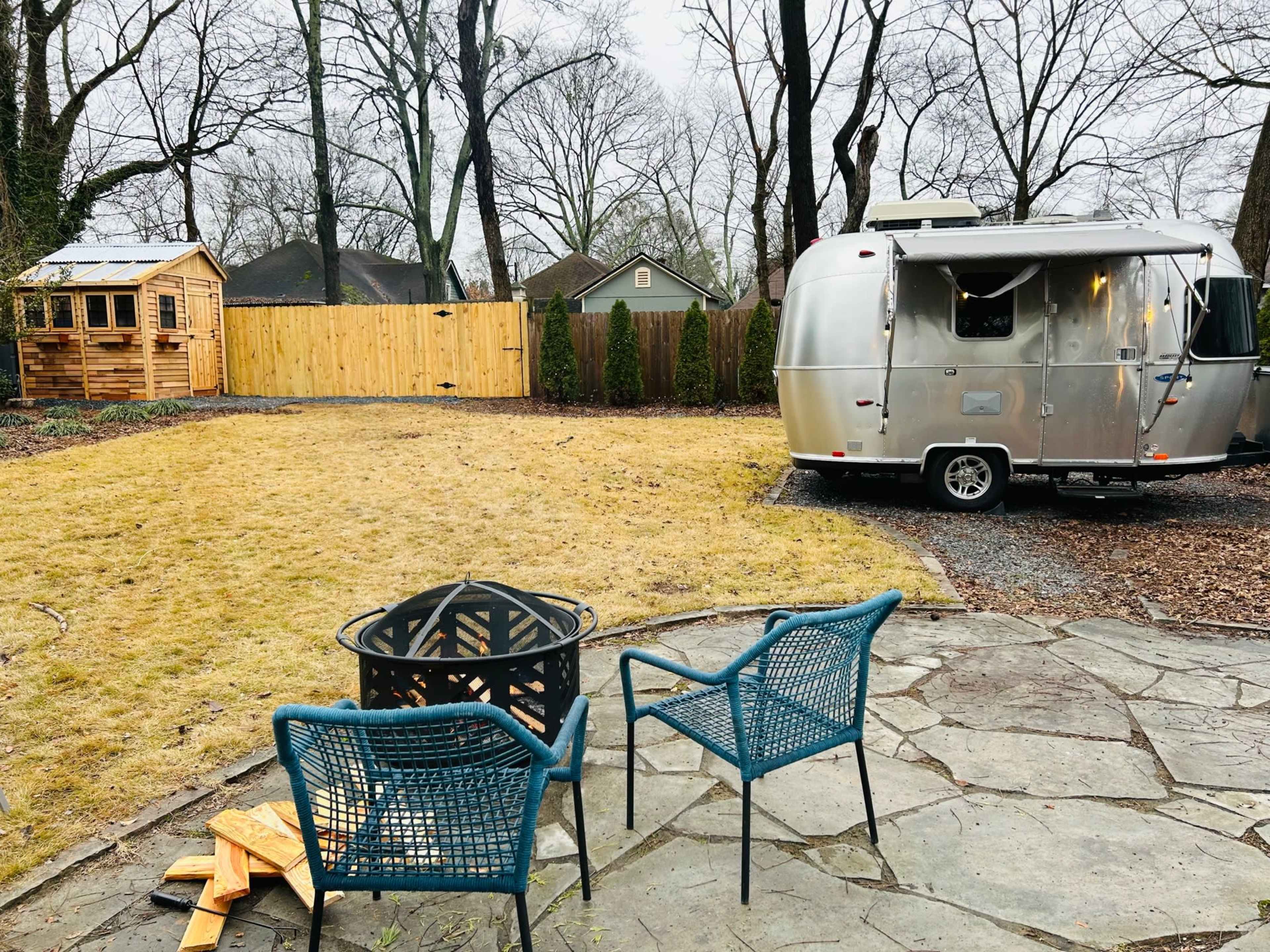 An Airstream trailer is parked beside a stone patio with two chairs and a fire pit, set in a yard with a wooden fence and a shed in the background.