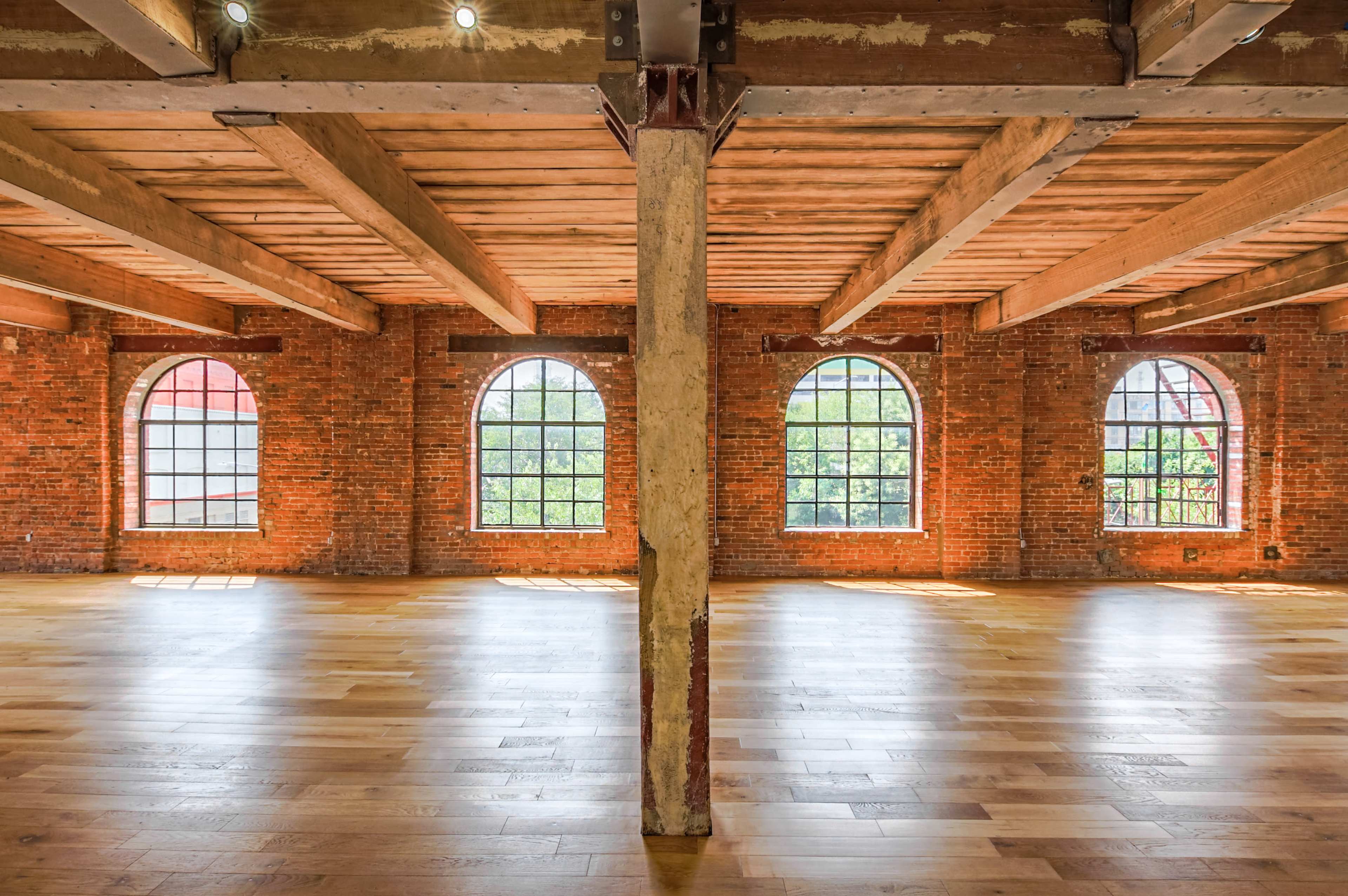 The image shows a spacious room with large arched windows, exposed brick walls, wooden beams, and a wooden floor.
