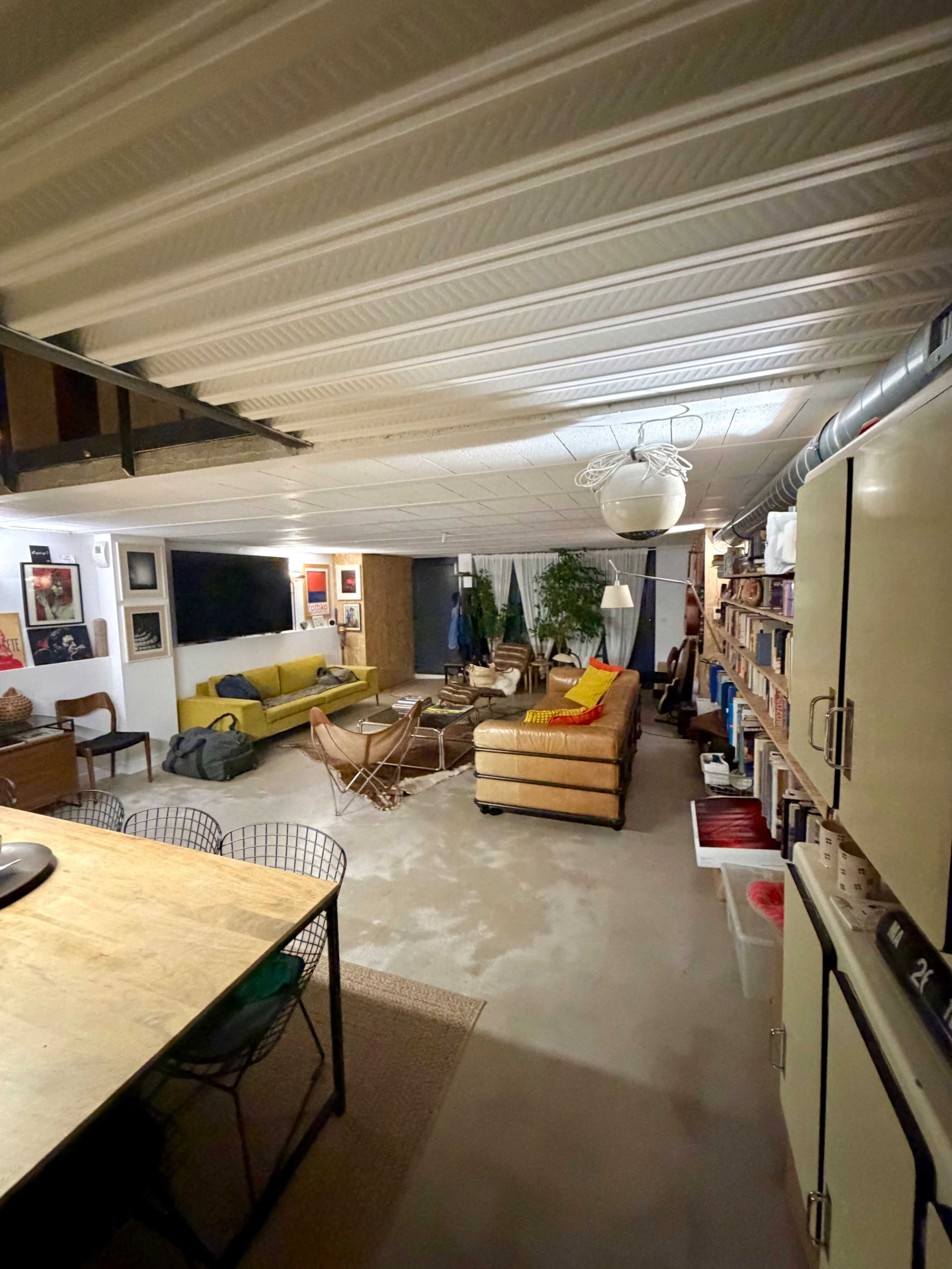 The image shows a spacious basement living area featuring a mix of seating options, including sofas and chairs, along with a dining table and shelves filled with books.
