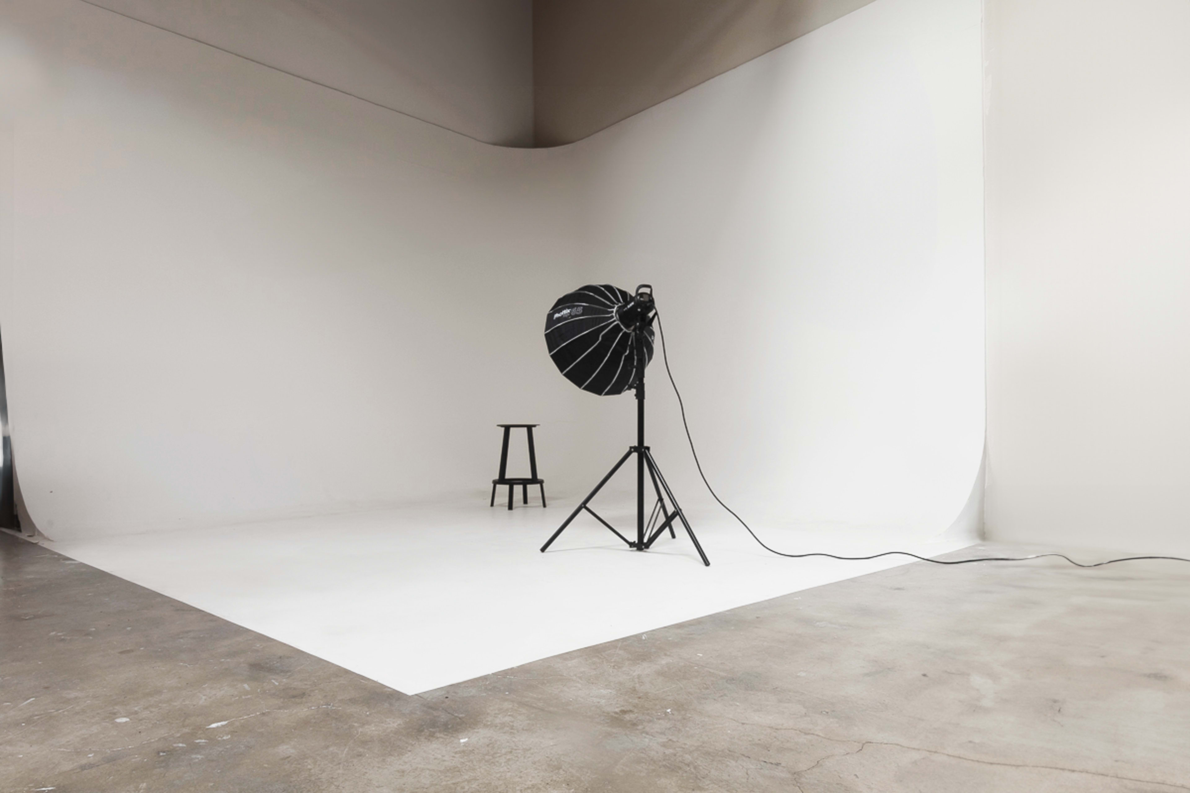 A photography studio features a single black light on a stand and a wooden stool against a white backdrop.