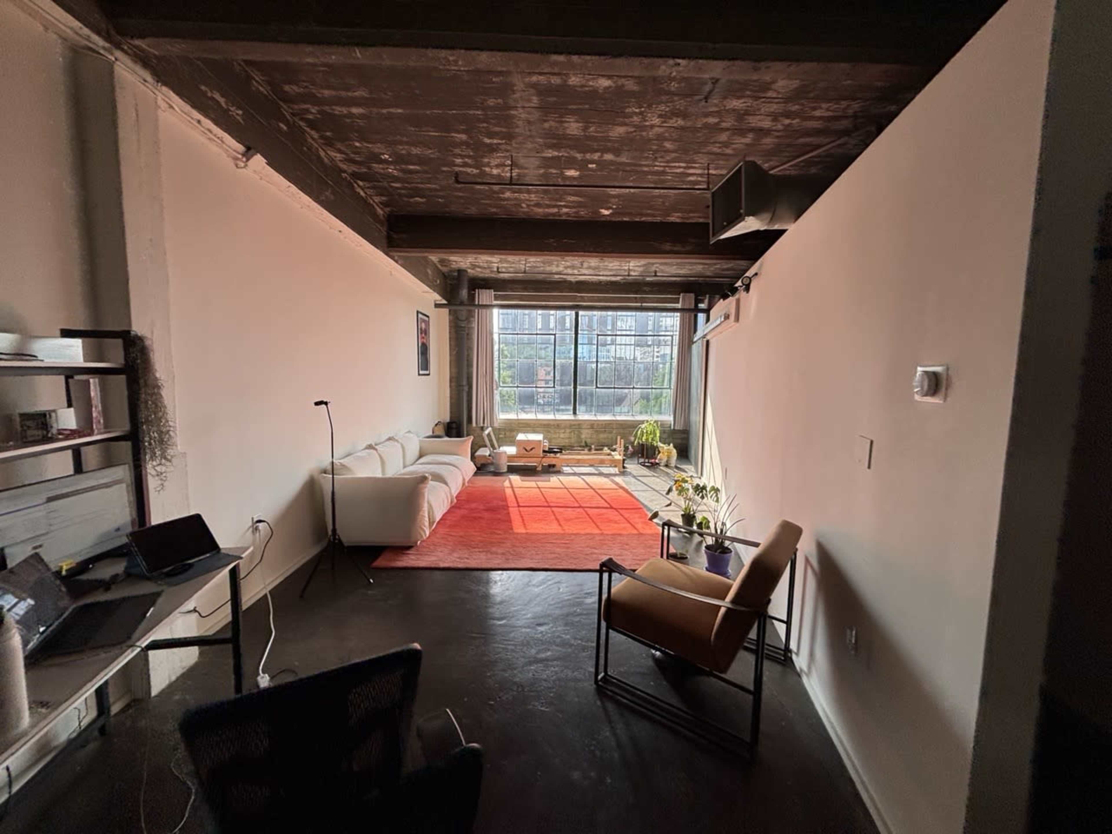 The image shows a spacious, minimalist living room with a large window, a white sofa, a red area rug, and a few plants.