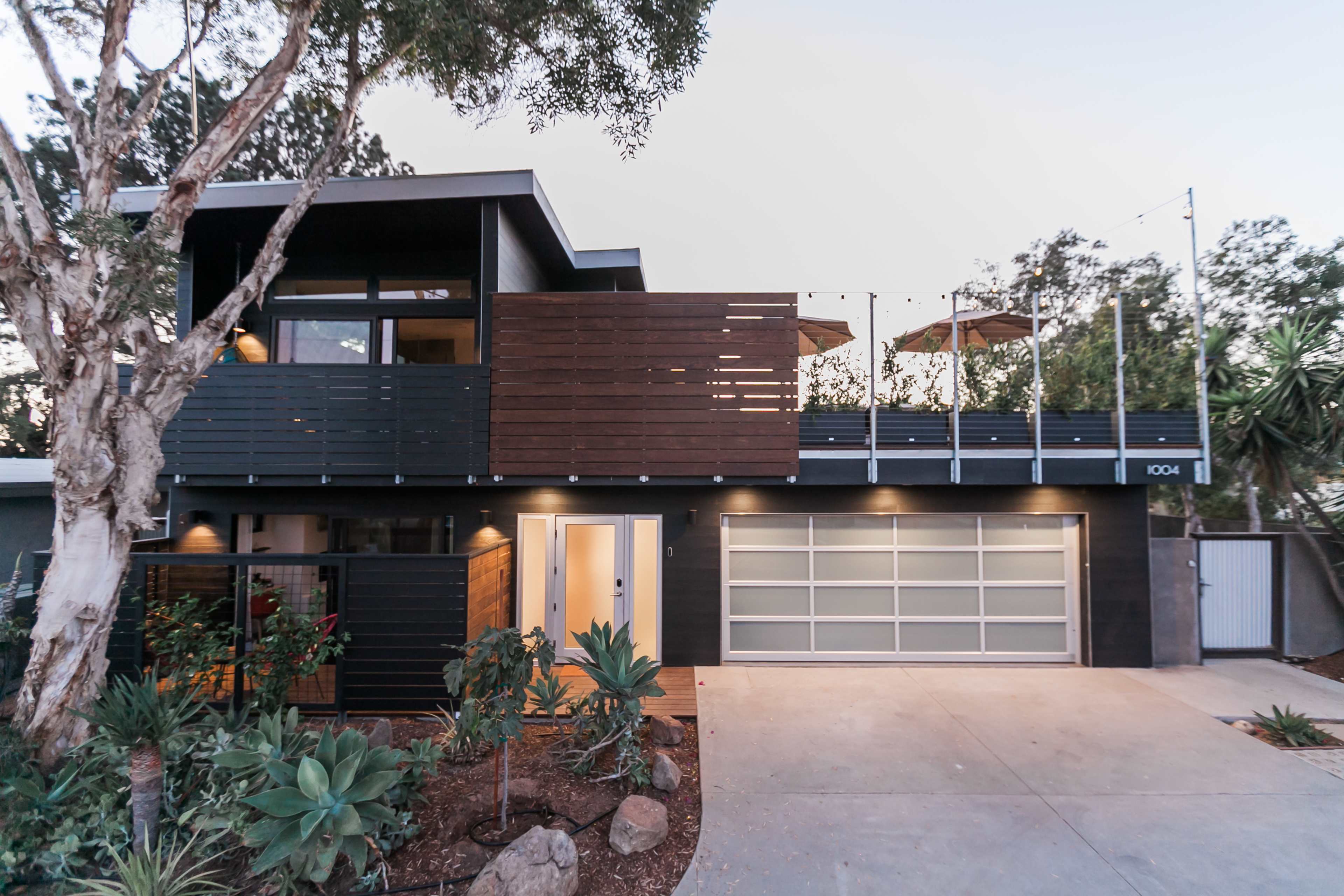 A modern two-story house with dark wood elements, a garage door, and landscaped greenery in front.