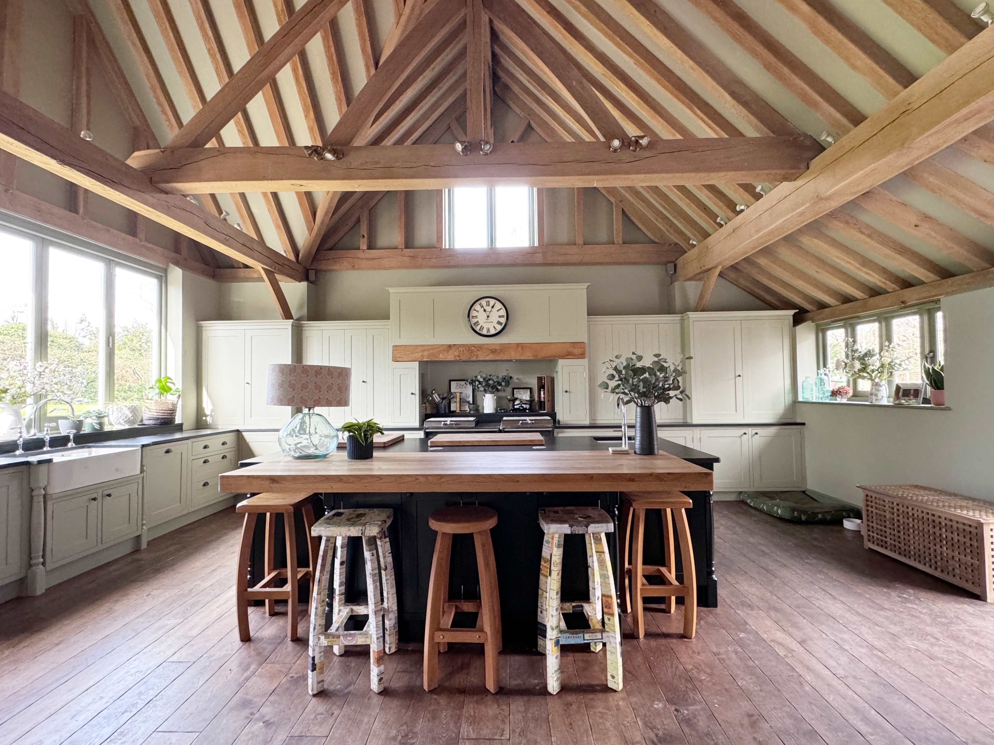 The kitchen features a large central island with wooden stools, exposed beams, and ample natural light from large windows.