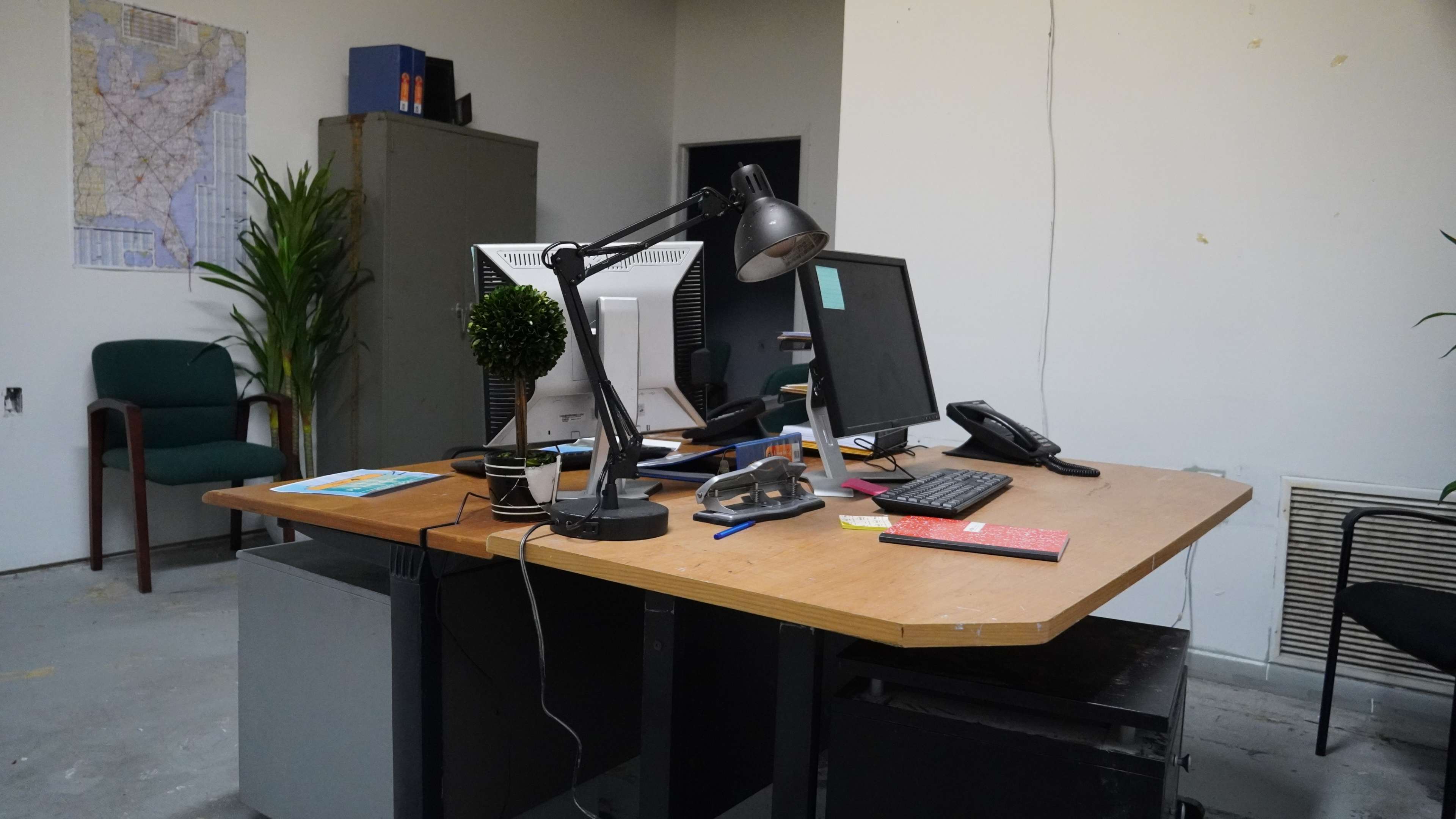 The image shows a sparsely furnished office with a desk featuring two computer monitors, a desk lamp, a telephone, and various office supplies, along with a map on the wall.