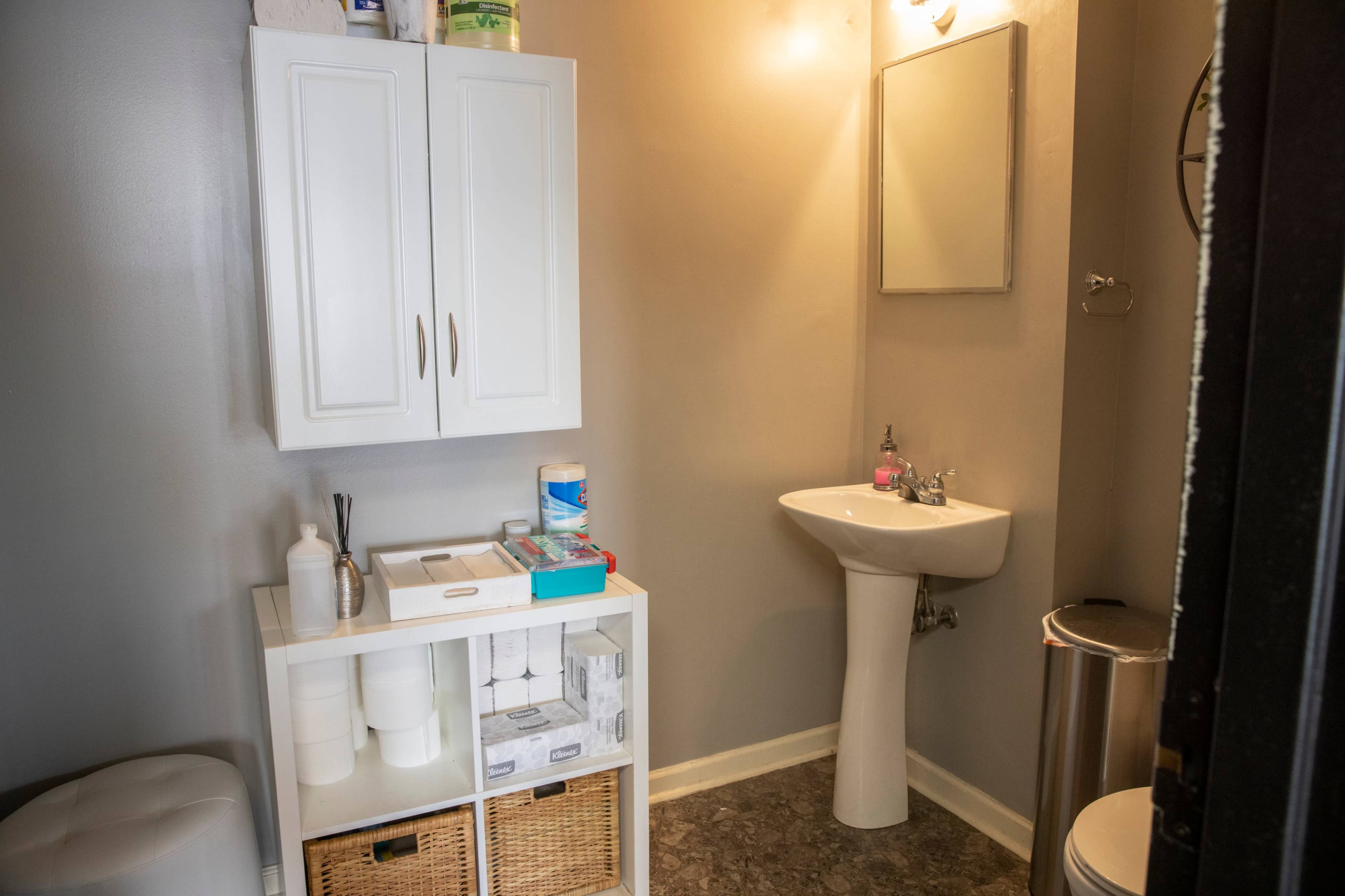A small bathroom featuring a sink with a mirror, a storage cabinet with shelves, and a waste bin.