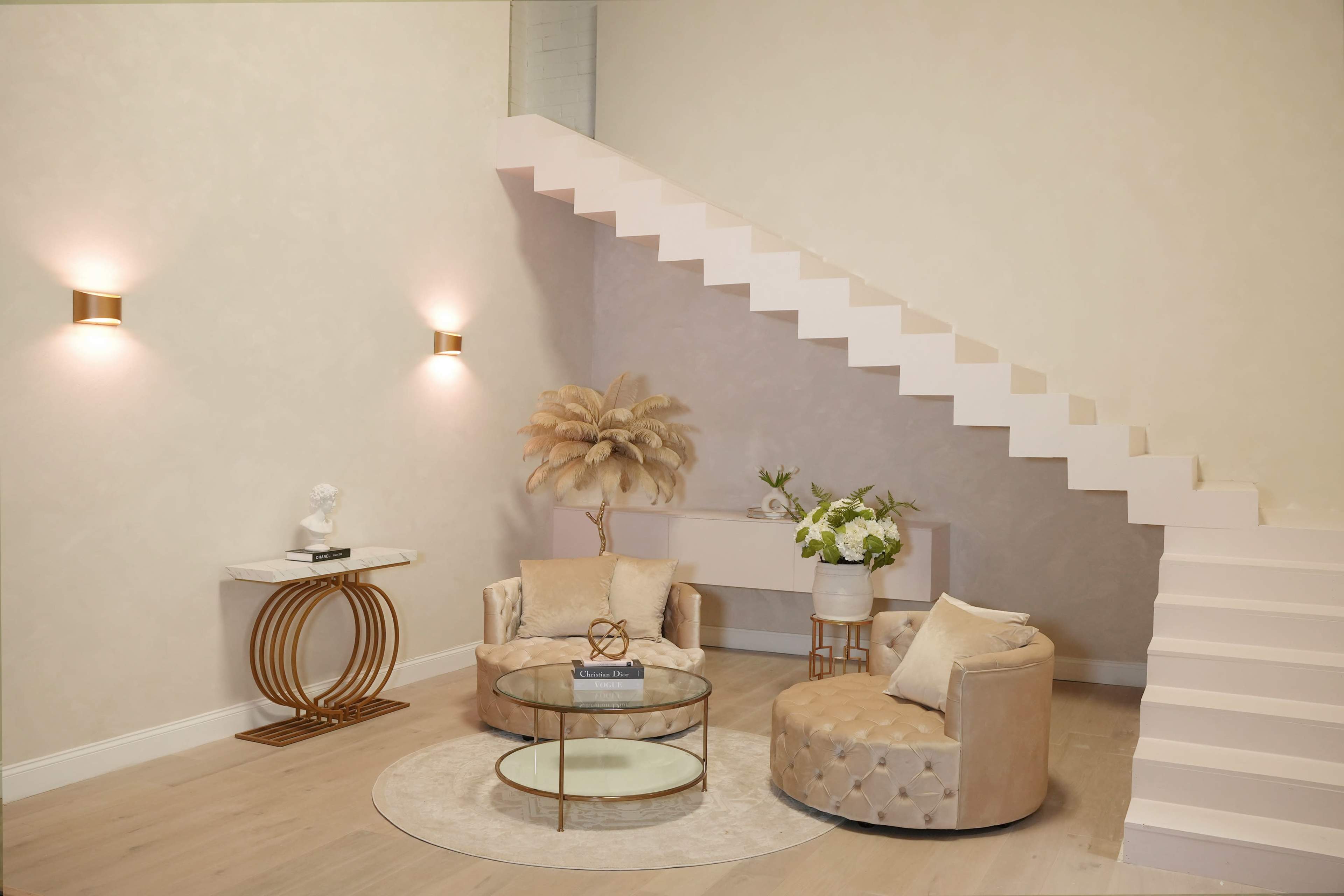 A modern living space with two plush armchairs, a round glass coffee table, and a staircase with minimalist design elements.