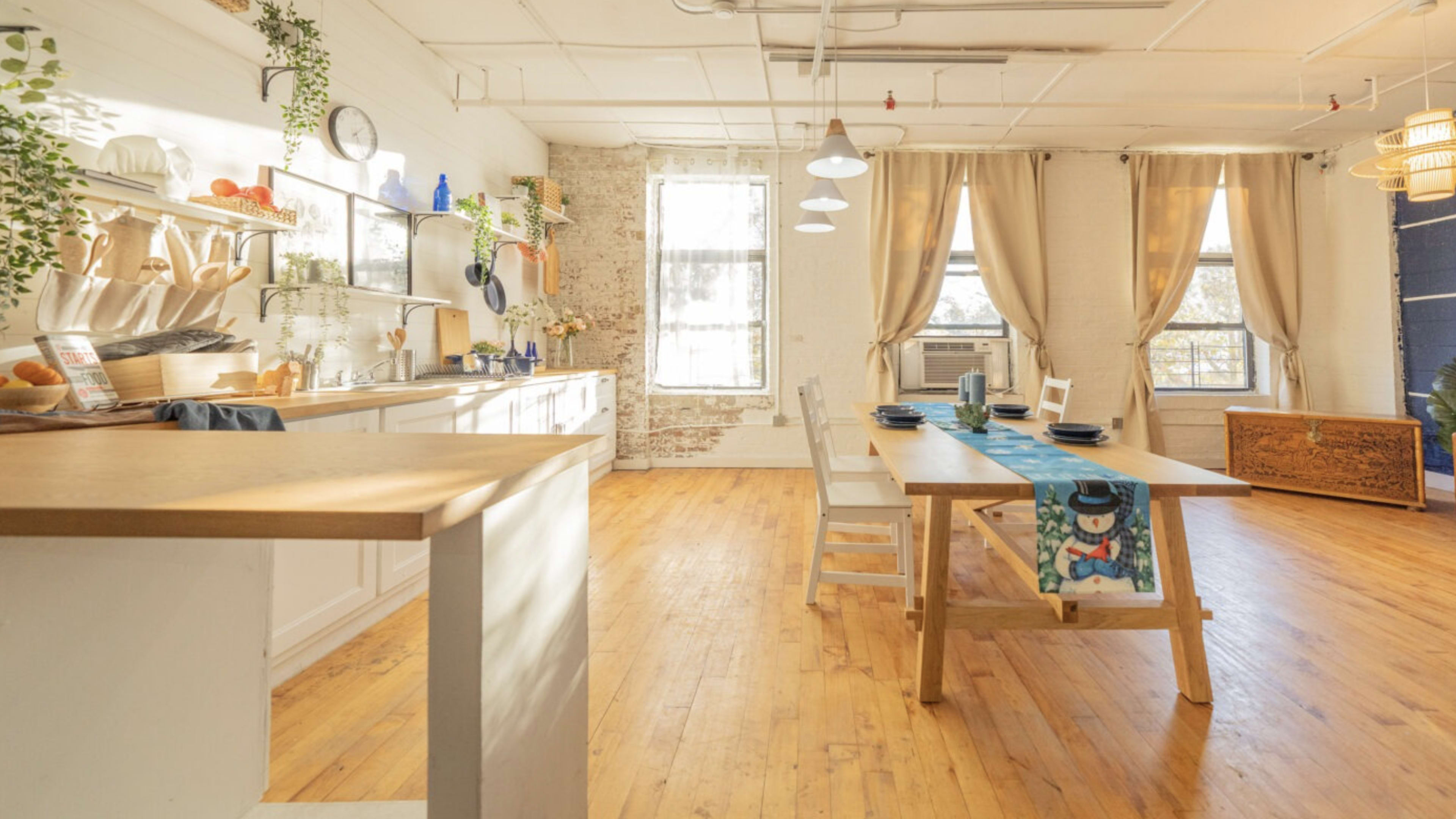 A bright and airy kitchen and dining area with a wooden table set for a meal, surrounded by large windows and decorated with plants.