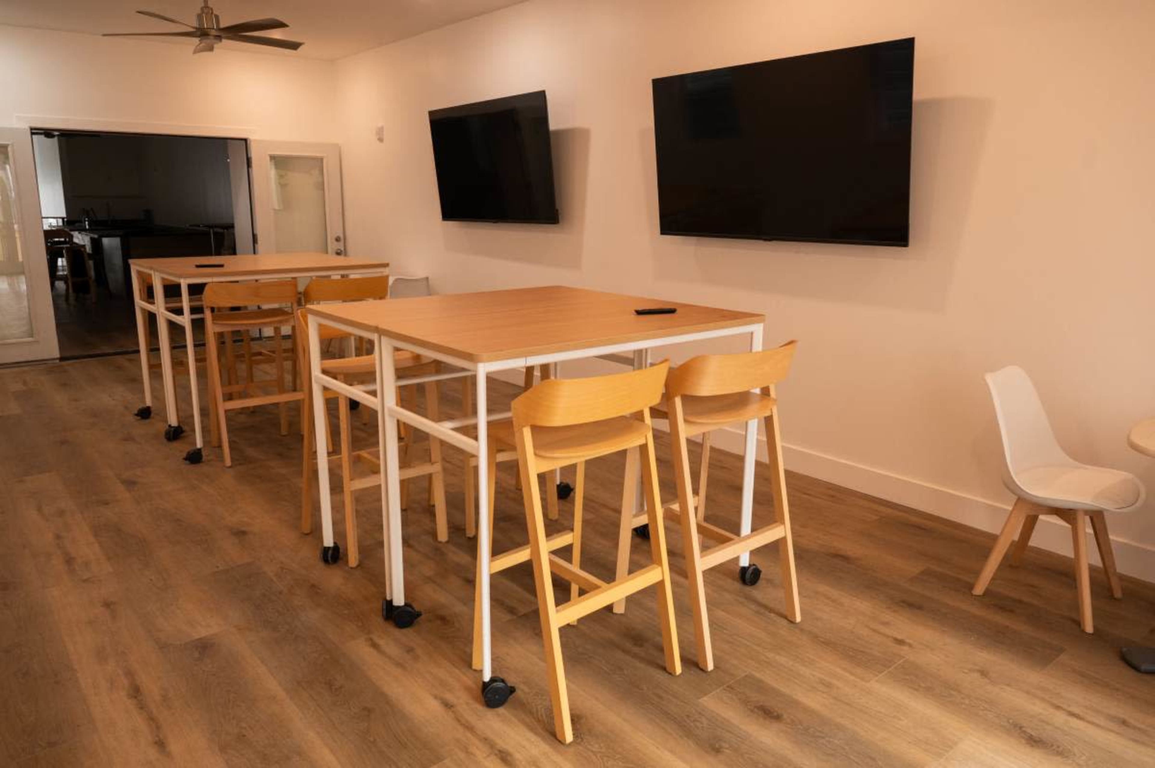 The image shows a modern workspace with multiple tall tables and chairs, two wall-mounted televisions, and wooden flooring.