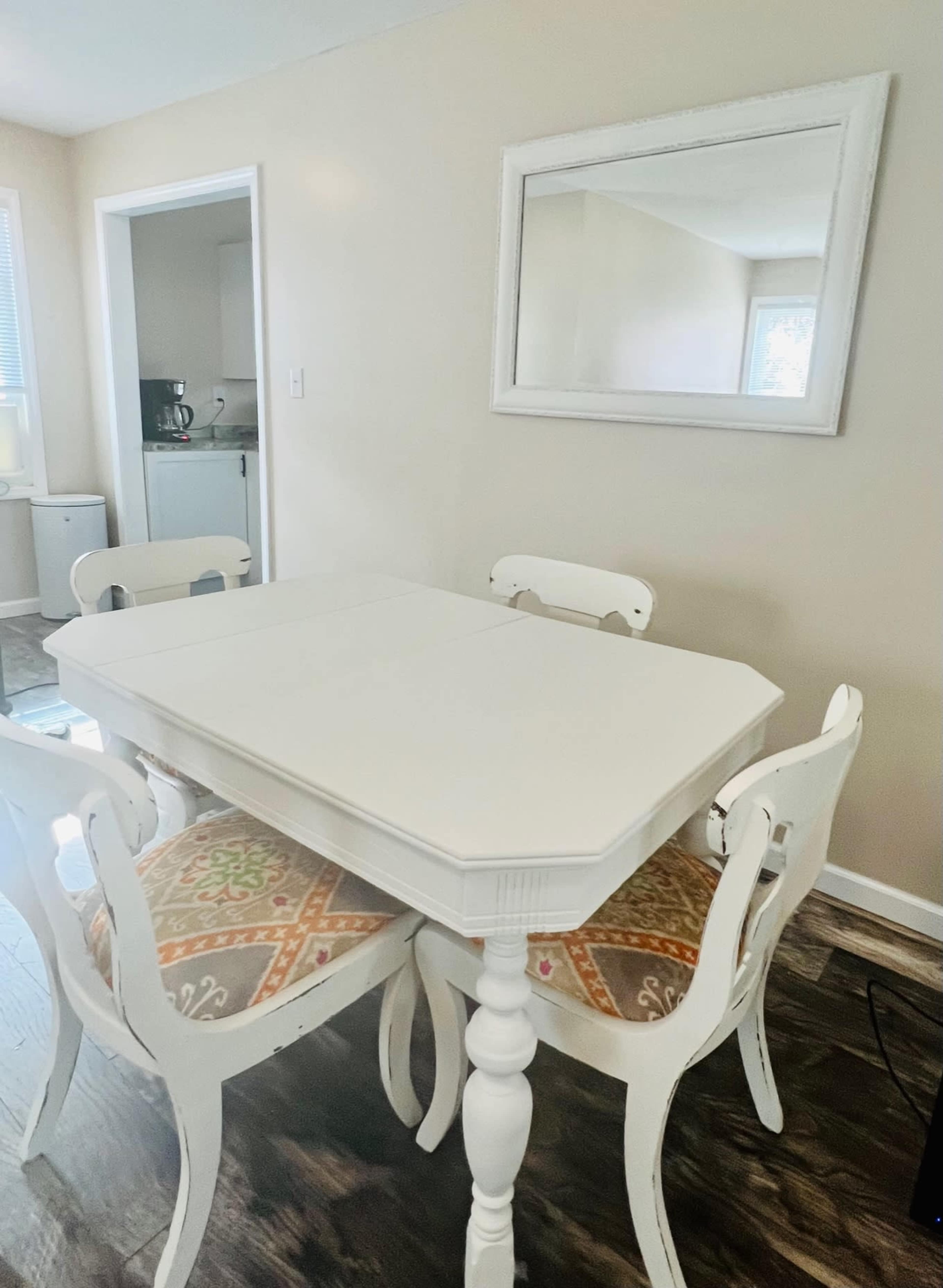 A white dining table with four matching chairs is set against a light-colored wall, with a mirror hanging above the table in a well-lit room.