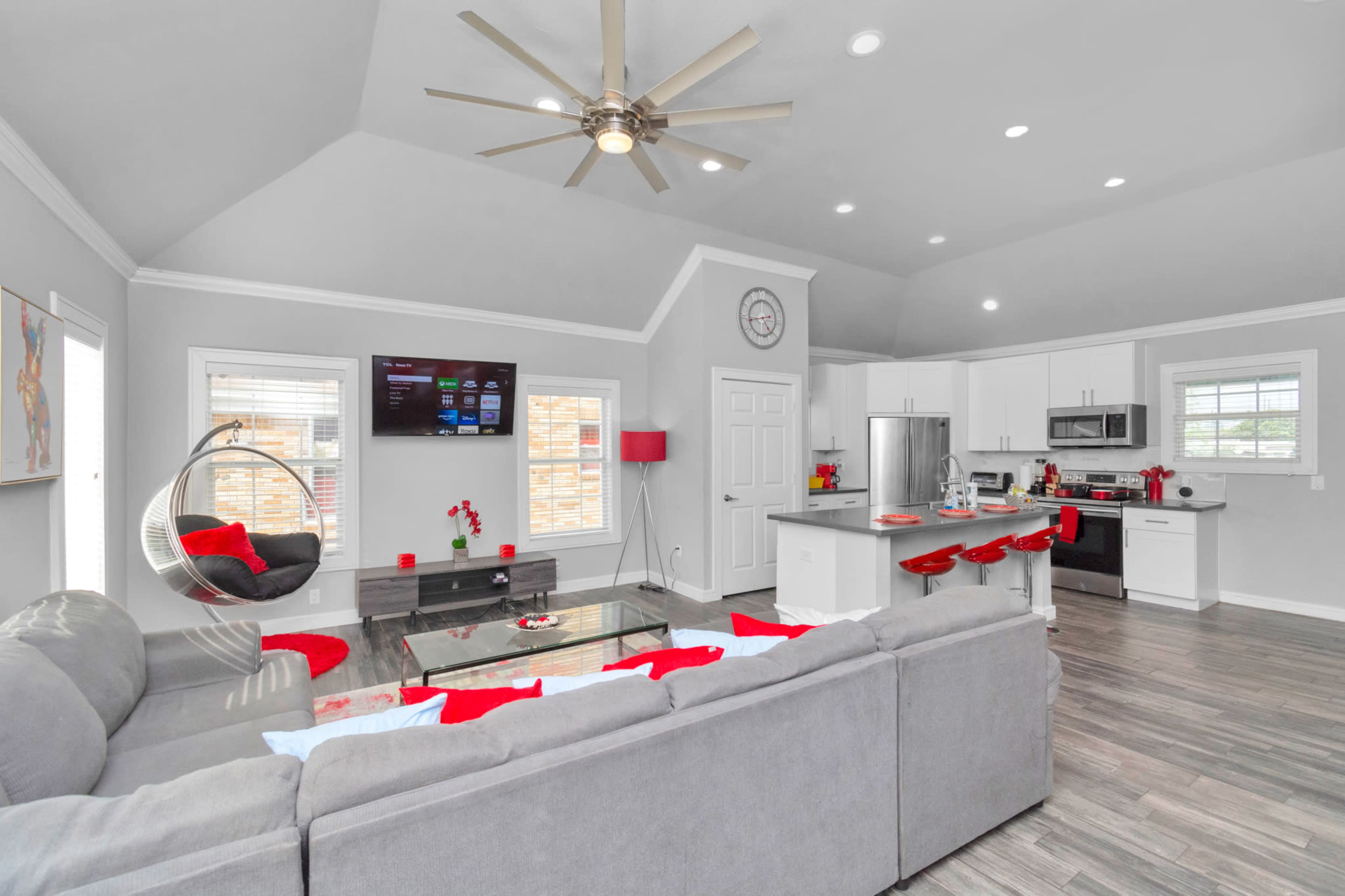 A modern living room features a gray sectional sofa, a glass coffee table, and a kitchen area with white cabinetry, all accented by red decorative elements.