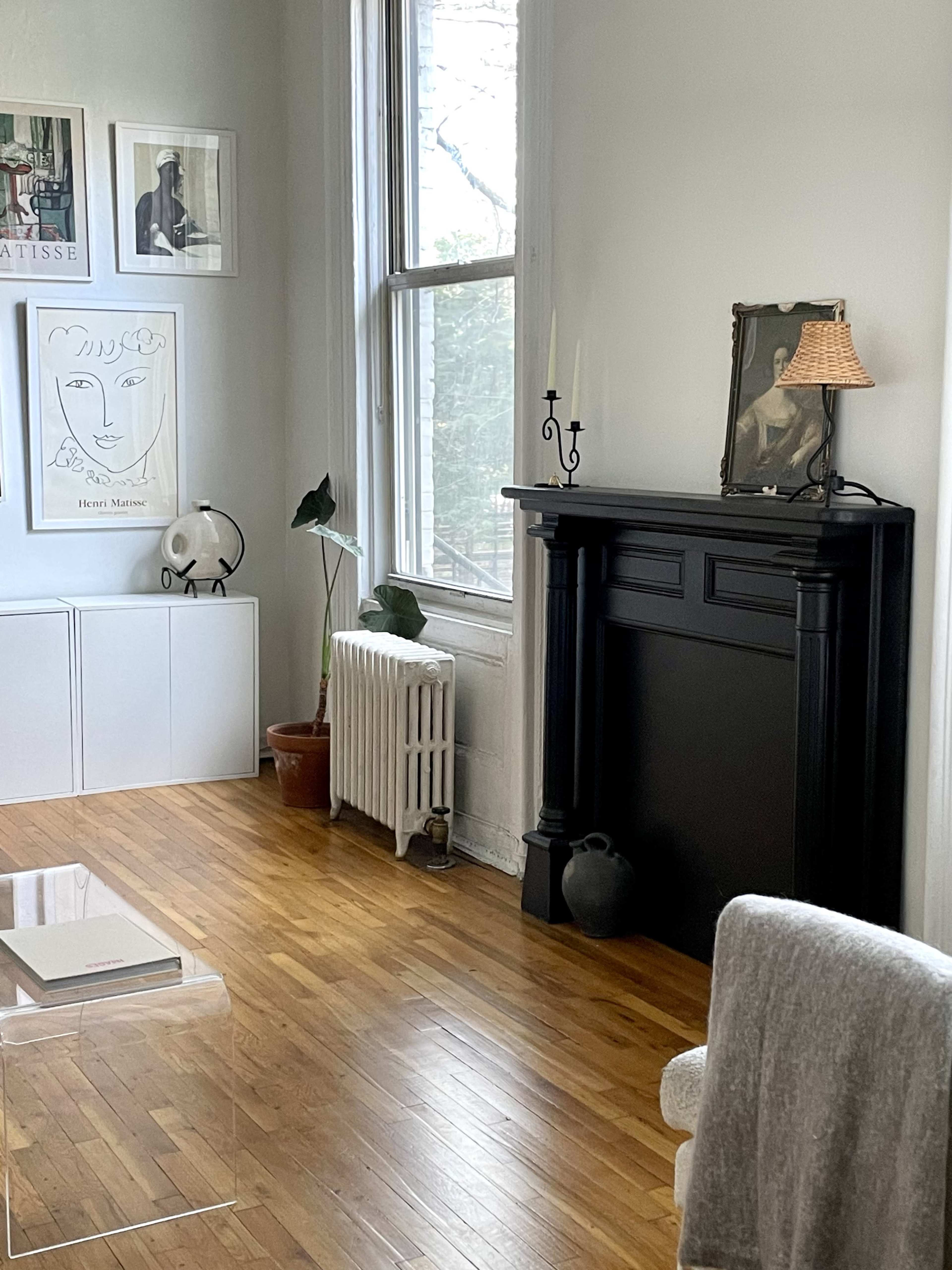 A minimalist living room features a black mantel, a radiator, a potted plant, and framed artwork on the light gray walls.