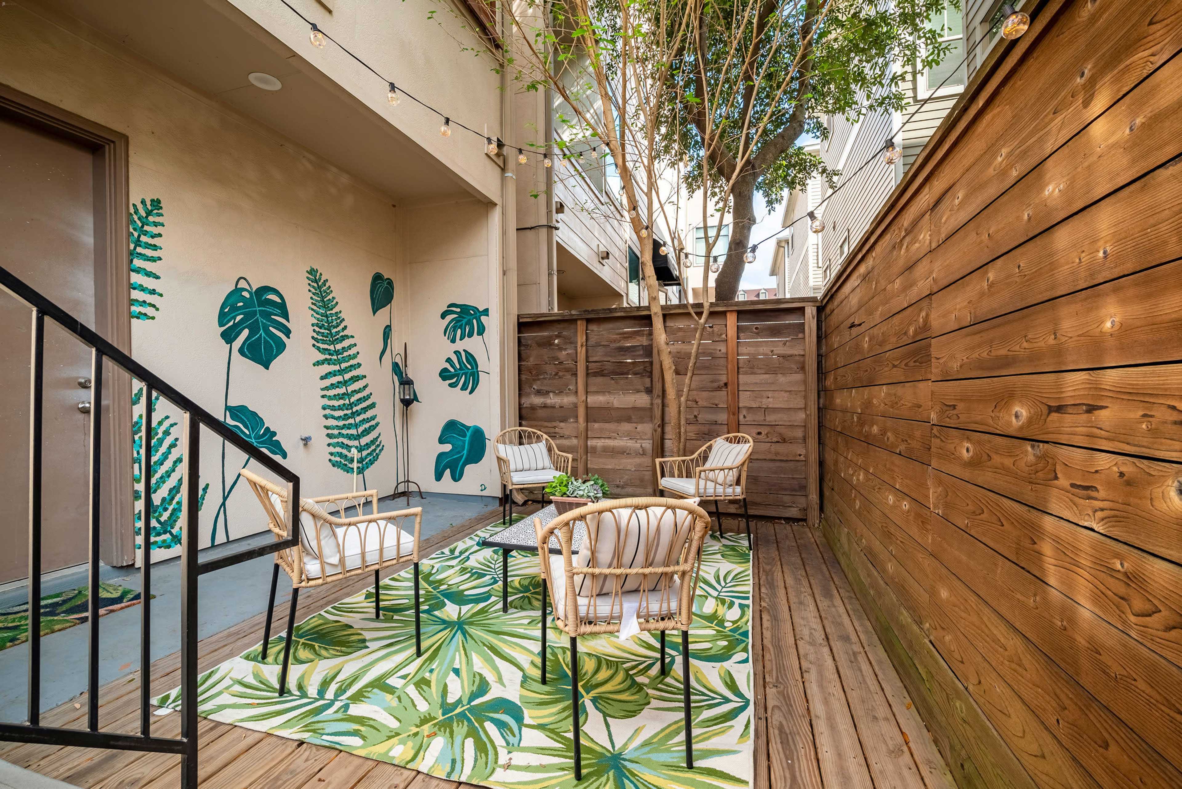 A cozy outdoor seating area features four chairs arranged around a table on a leaf-patterned rug, surrounded by wooden walls and decorative greenery.