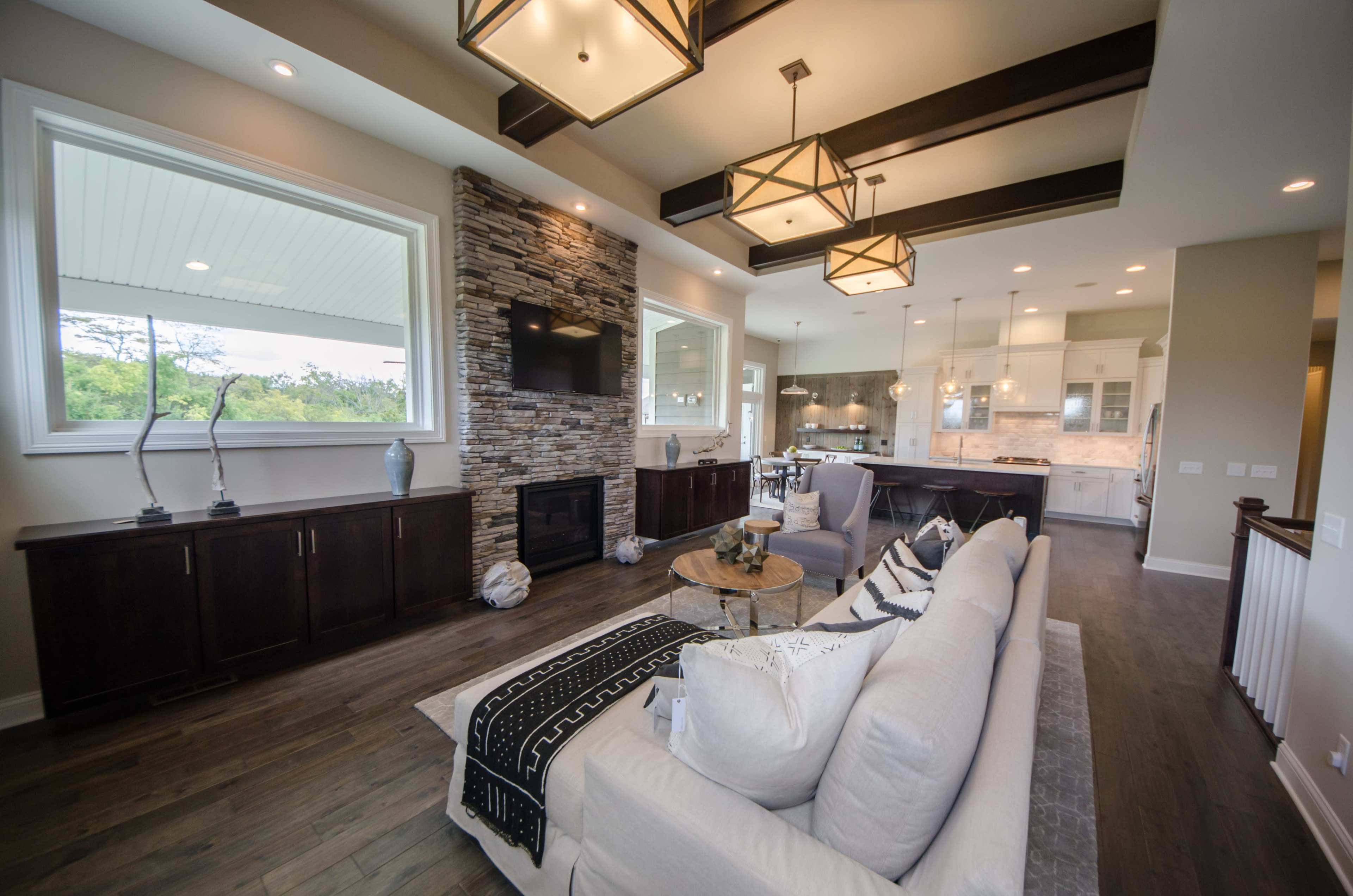 A modern living room with wooden floors, a stone fireplace, large windows, and a sectional sofa.
